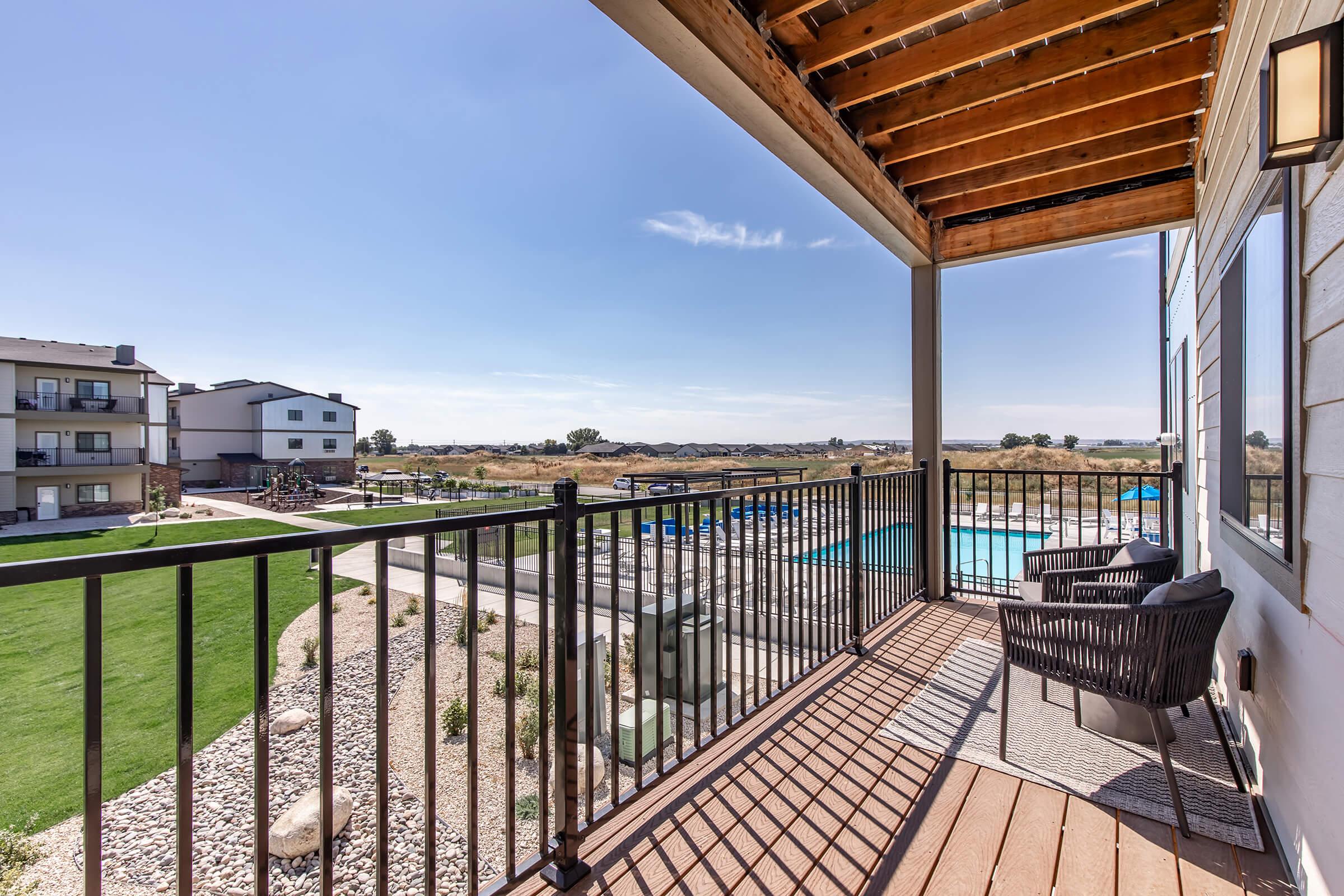 View from a balcony overlooking a pool and landscaped area. Features two black chairs and a small rug, with a backdrop of buildings and an open grassy space under a clear blue sky. The scene captures a peaceful outdoor setting with a mix of residential and recreational elements.