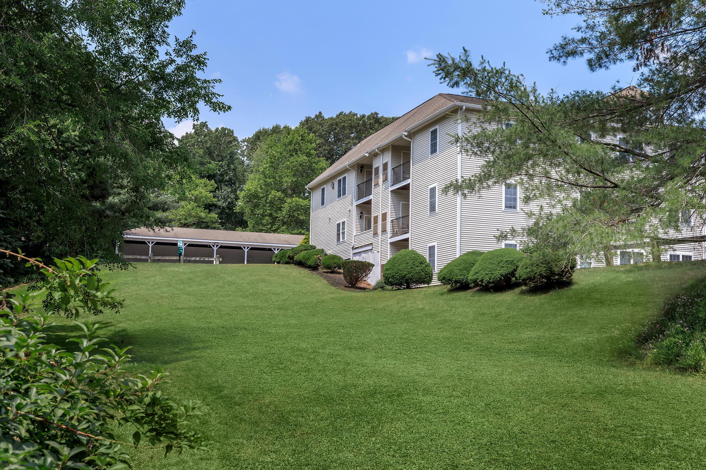 Apartments in Manchester CT - The Manchester - Apartment Building Exterior With Balconies, Manicured Green Lawn, Mature Trees, And Covered Parking In A Quiet Setting