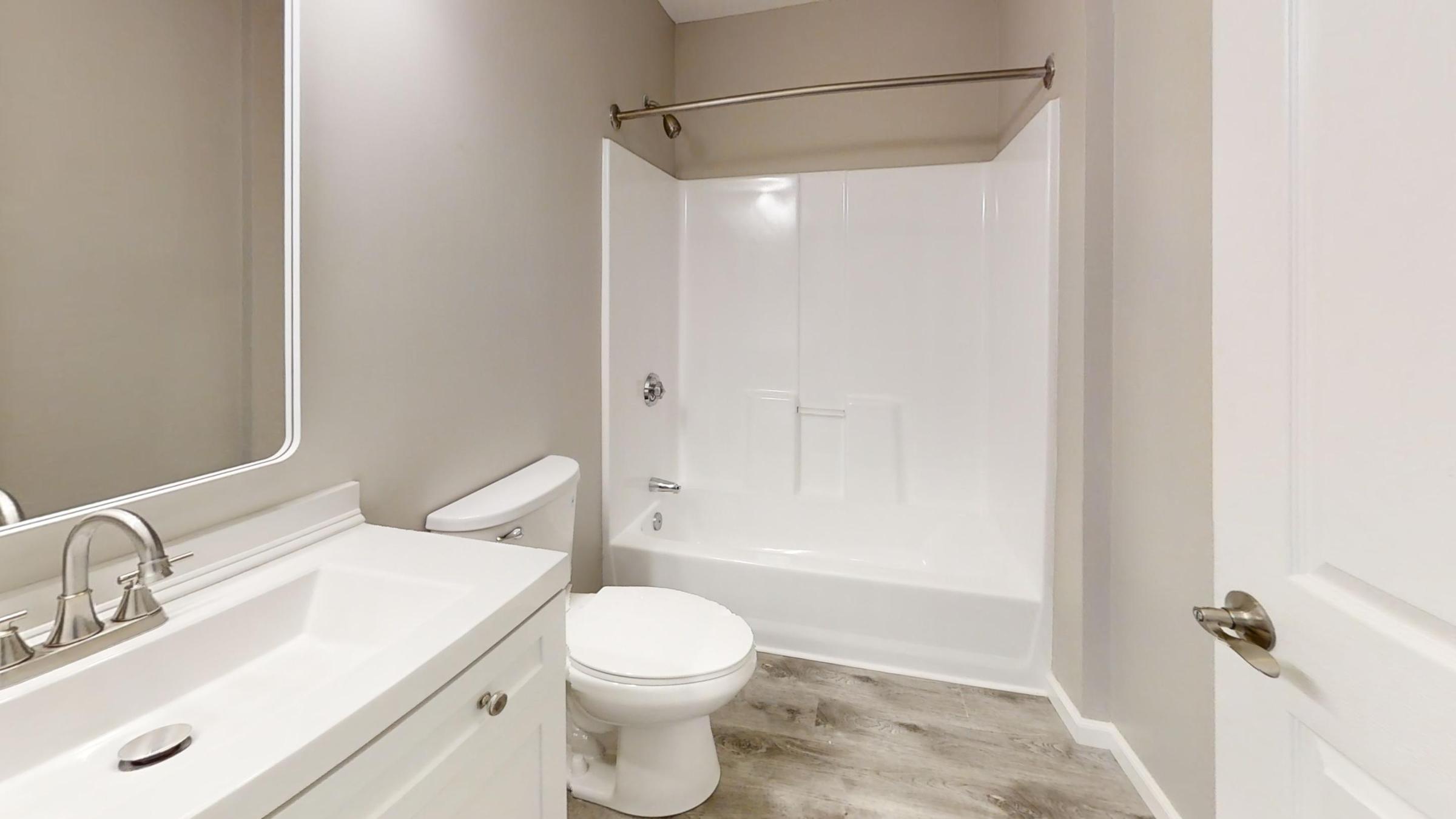 A modern bathroom featuring a white bathtub and shower combination, a white toilet, and a sleek vanity with a sink. The walls are painted a light beige, and the floor is covered with wood-like tiles. A large mirror above the sink enhances the bright and spacious feel of the room.