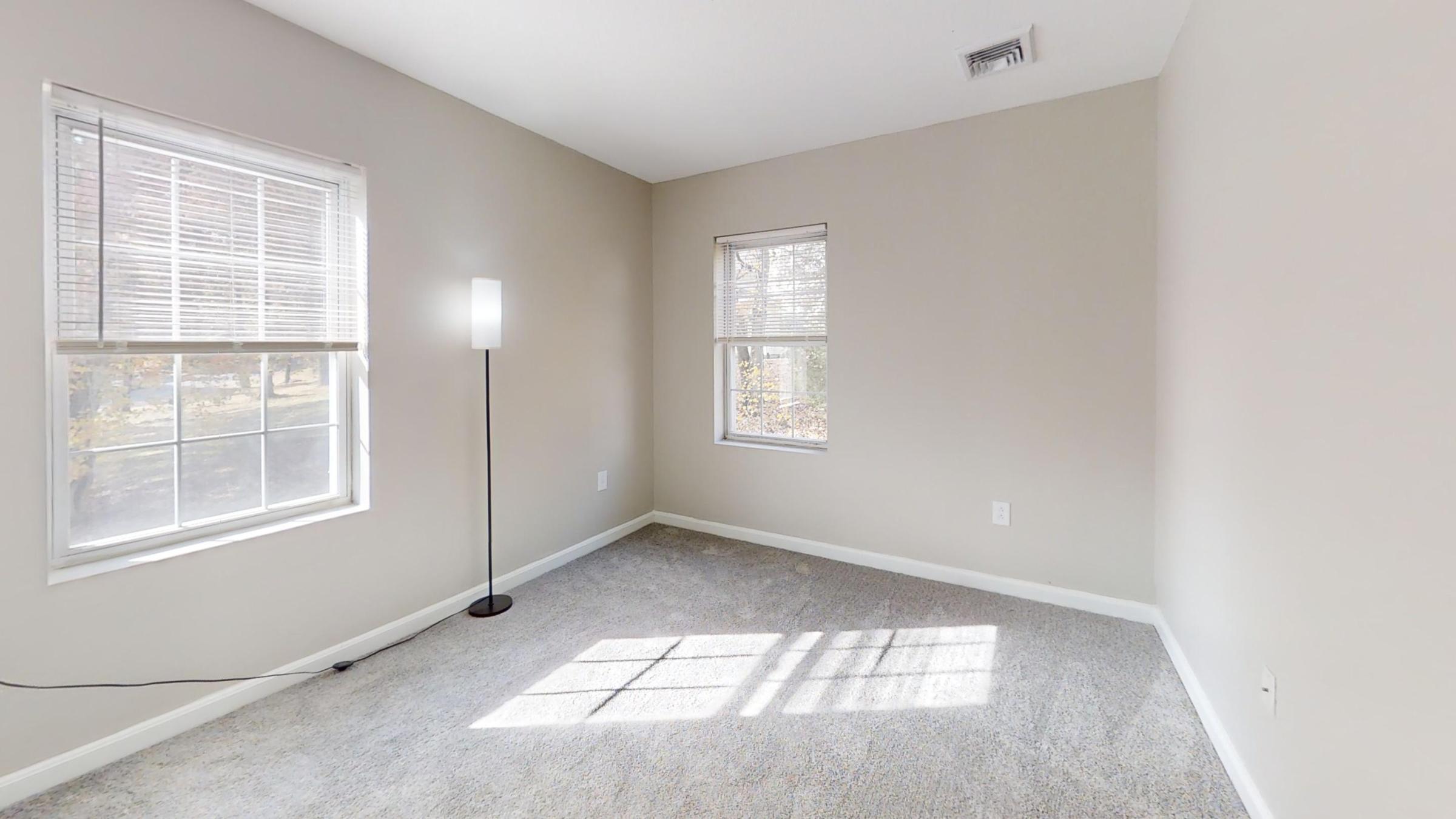 A minimalistic room featuring soft beige walls and light carpeting. Two windows allow natural light to enter, casting shadows on the floor. A slender floor lamp stands in the corner, enhancing the bright and airy atmosphere of the space. The room appears empty and ready for furnishing.