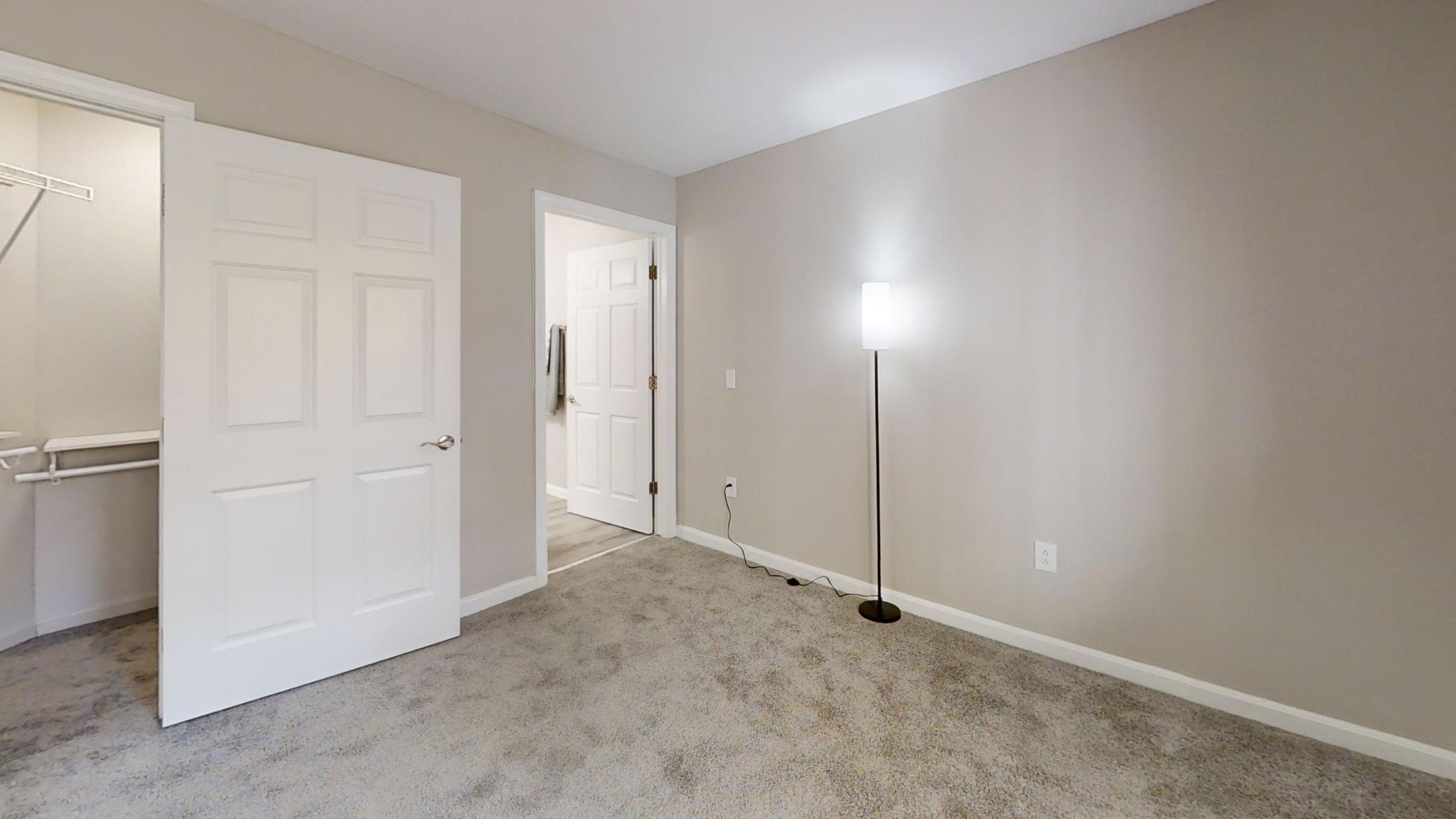 Empty room with beige walls and carpeted floor. There is a white lamp on a black stand in the corner. A closed door leads to a closet on the left, while an open door on the right leads to another room. The lighting is soft, creating a neutral and inviting atmosphere.