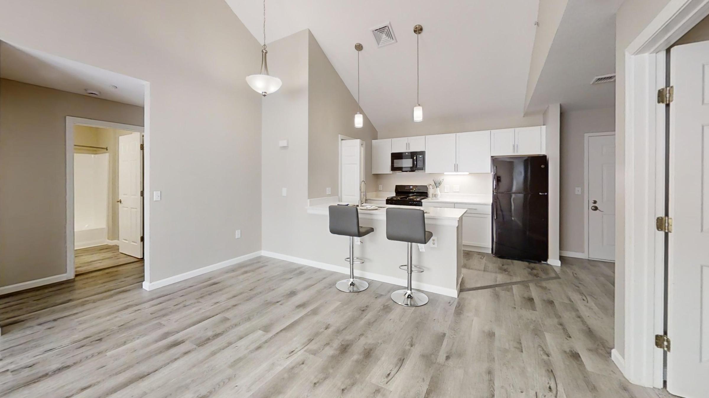 A modern kitchen and dining area featuring a white cabinetry, a black stove, and refrigerator. Two bar stools are positioned at the kitchen island. The room has light-colored walls and flooring, with a doorway leading to another room in the background. Bright, natural light fills the space.