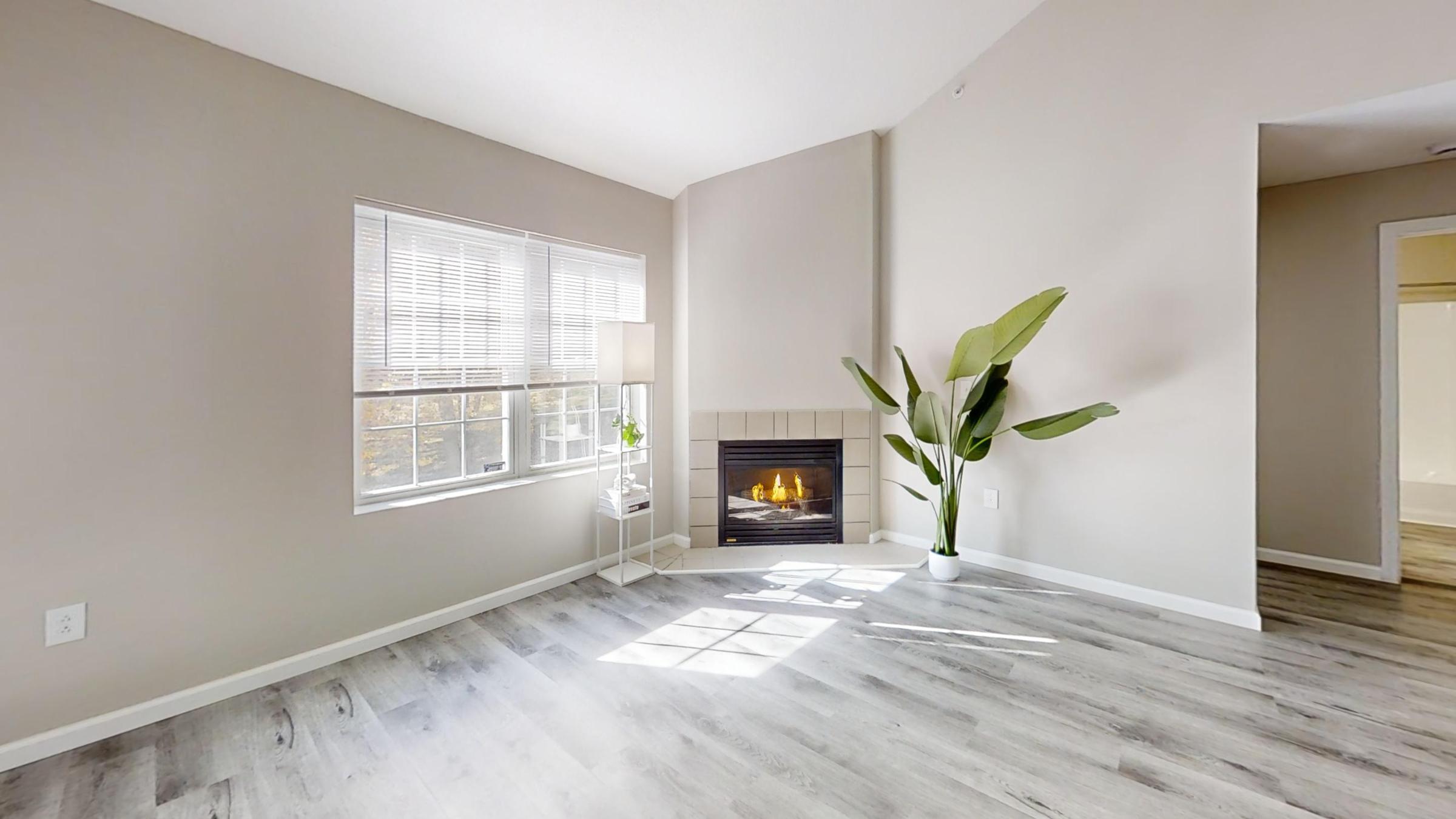 A bright and airy living room featuring light gray walls and wood flooring. A modern fireplace is positioned in the corner, accompanied by a floor lamp. A large window allows natural light to fill the space, and a tall green plant adds a touch of life to the room.