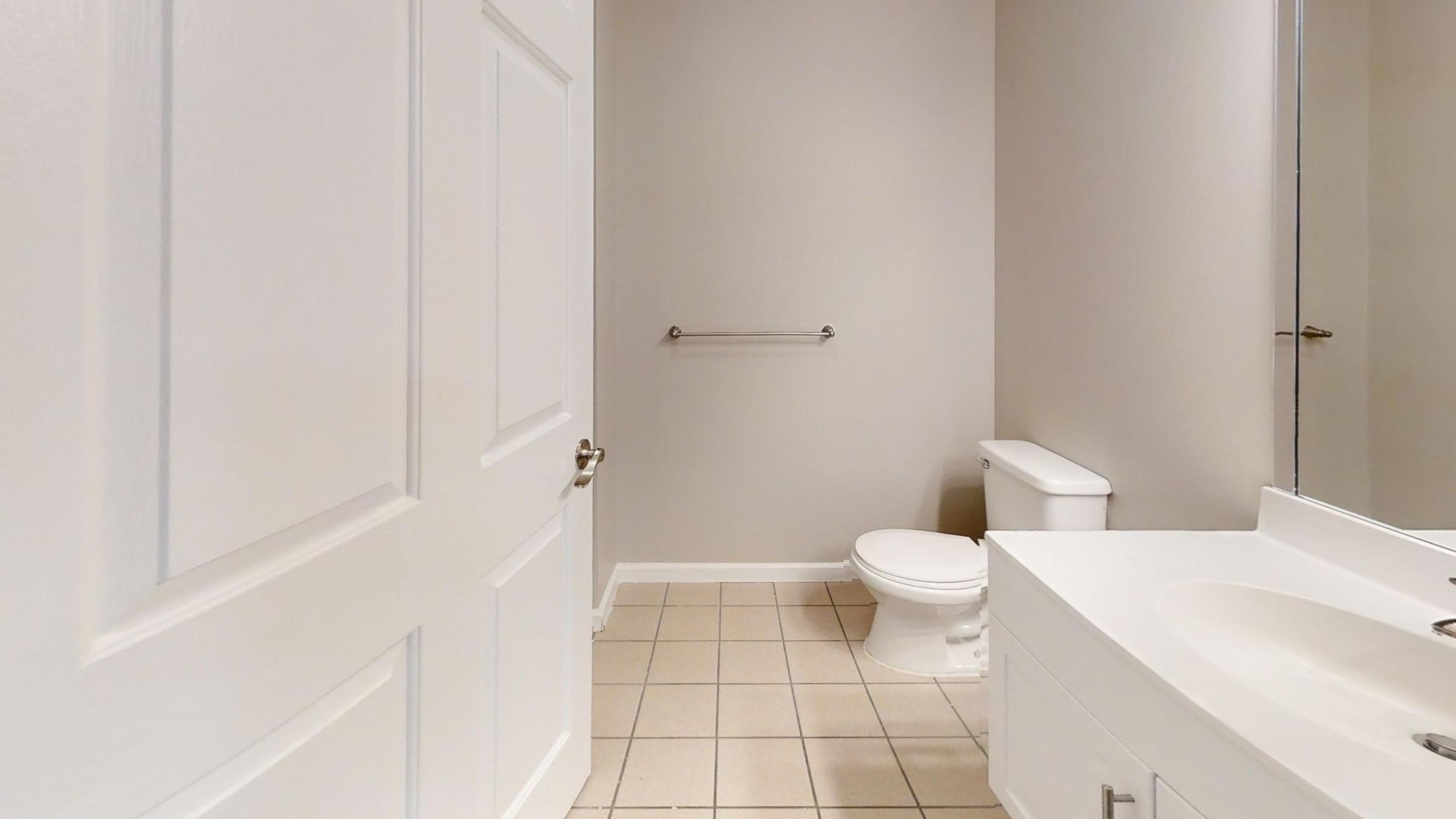 A clean, modern bathroom featuring a white toilet, a sleek vanity with a sink, and a large mirror. The walls are painted in a neutral color, and the floor is tiled. A towel bar is mounted on the wall, and there is a door leading out of the space. The overall ambiance is bright and uncluttered.