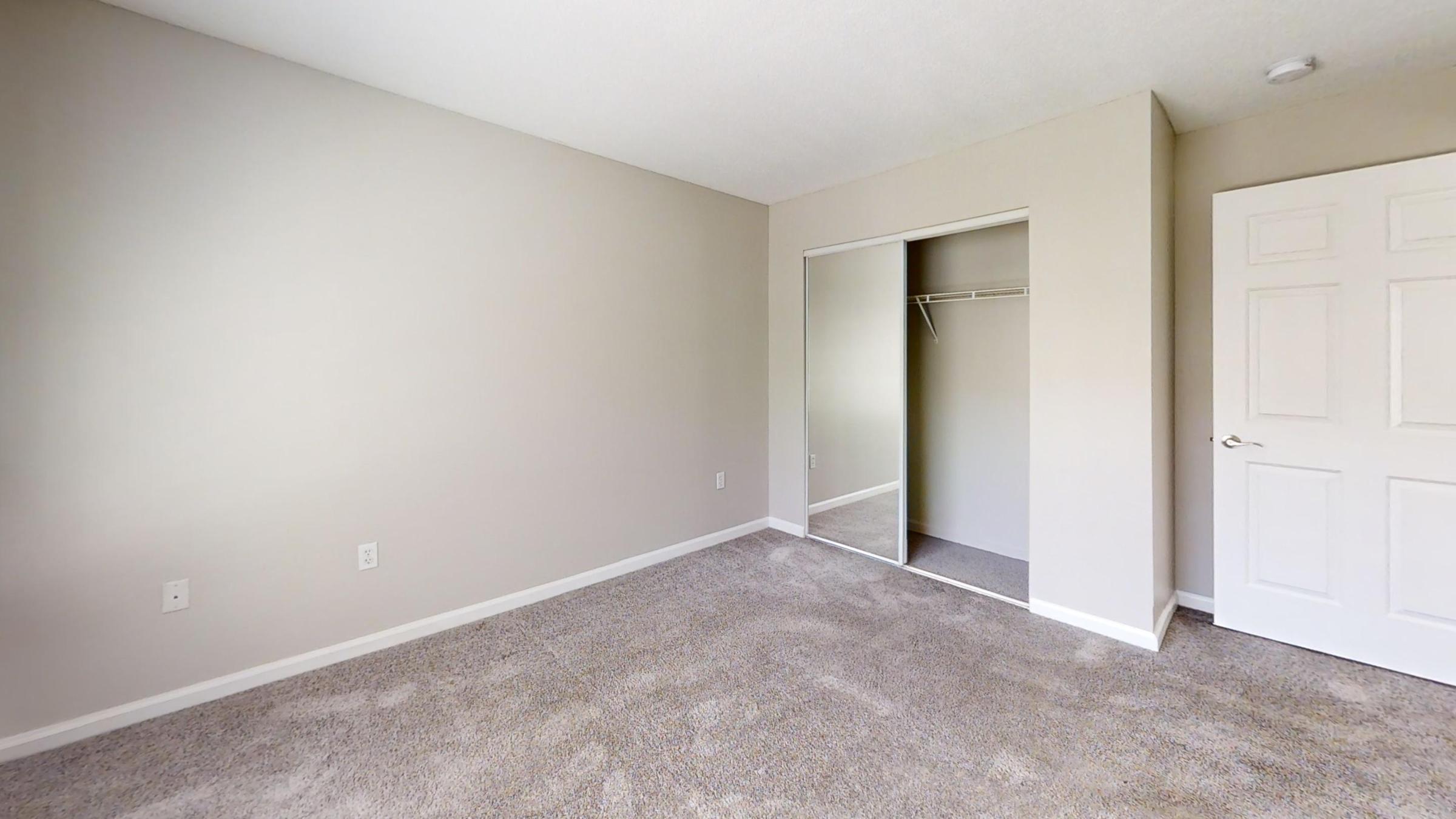 A sparsely furnished room featuring light-colored walls, a carpeted floor, and a sliding mirrored closet door. There are no furniture items present, creating an open and minimalist space. The room is well-lit, with natural light apparent from an unseen window.