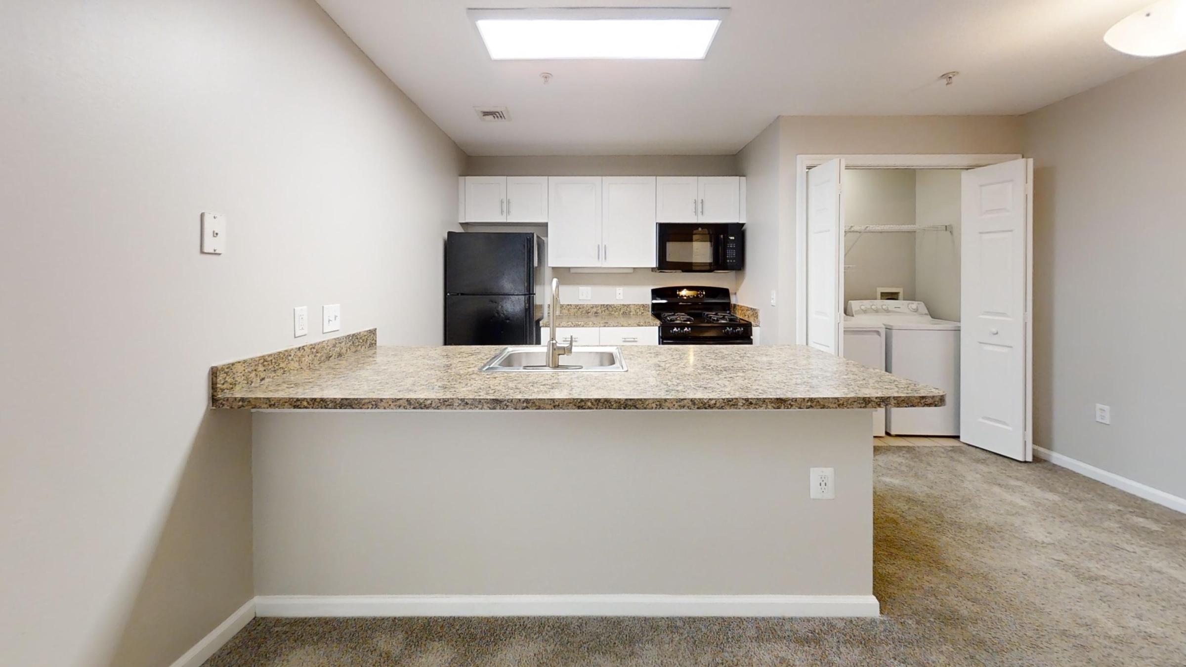 Spacious kitchen with a granite countertop, featuring white cabinets, a black microwave, and refrigerator. A laundry area is visible through an open door. The room has neutral-colored walls and carpet flooring, illuminated by overhead lighting, creating a modern and inviting atmosphere.