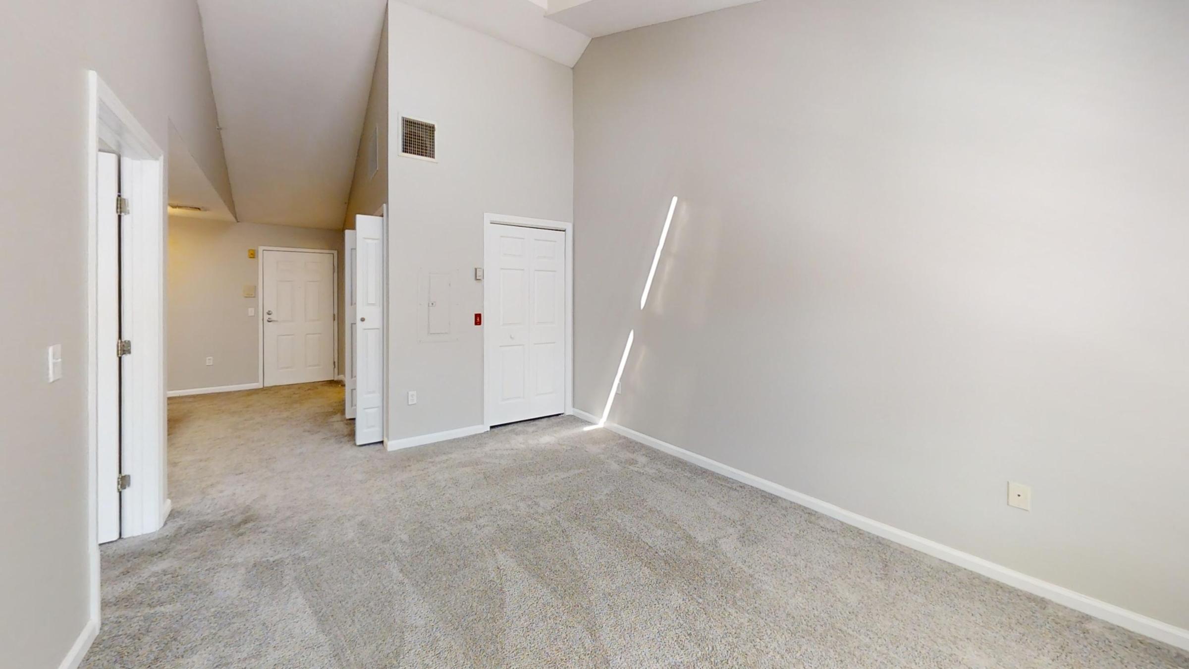 A spacious, empty room with light gray walls and plush carpet. Two closed doors are visible on the left, leading to other areas of the space. Natural light streams in from a window, creating a bright atmosphere. The ceiling is high, enhancing the open feel of the room.