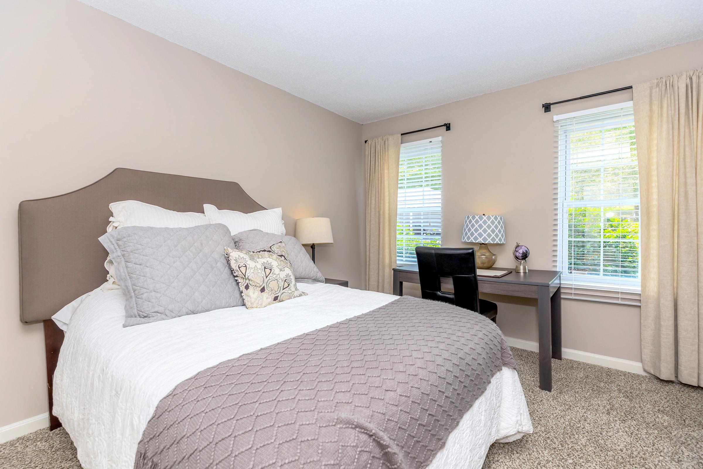 Apartments for Rent in Manchester CT - The Manchester - Bright Bedroom With Queen Bed, Gray Quilted Bedding, Desk Workspace, Soft Carpet, And Large Windows With Curtains