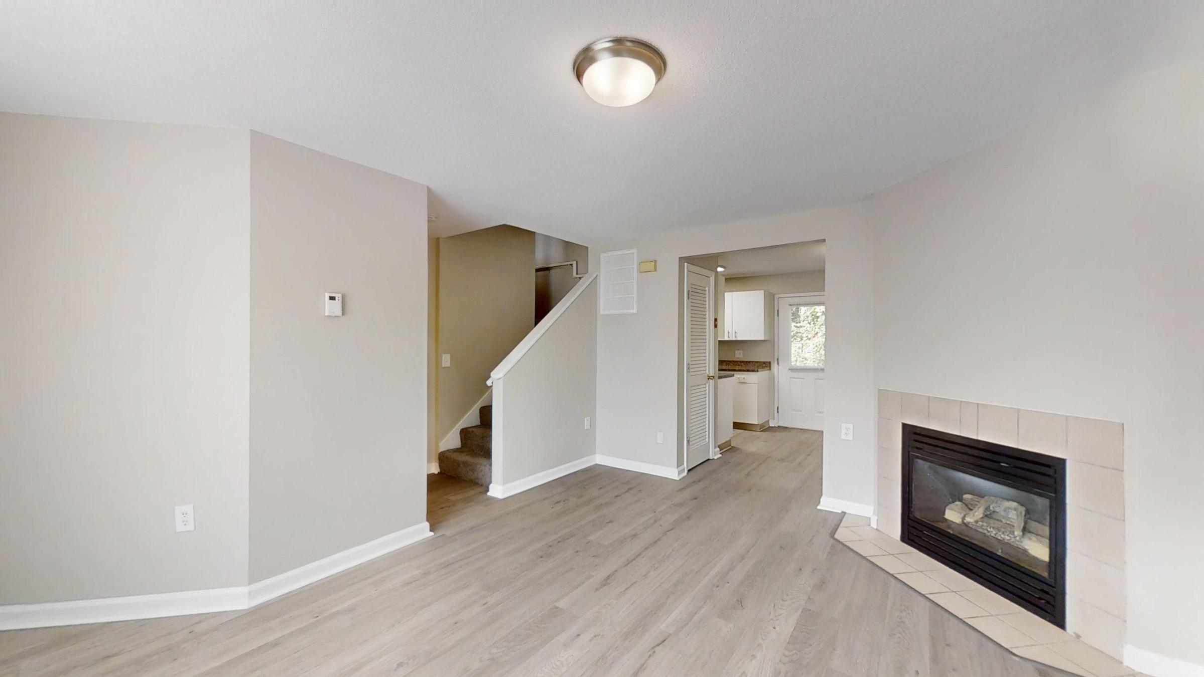 Cozy interior of a living space featuring light-colored walls, a modern light fixture, and a gas fireplace. A staircase is visible on the left, leading to the upper level, while an open doorway provides a view into a kitchen area. The flooring is a warm wood tone, enhancing the inviting atmosphere.