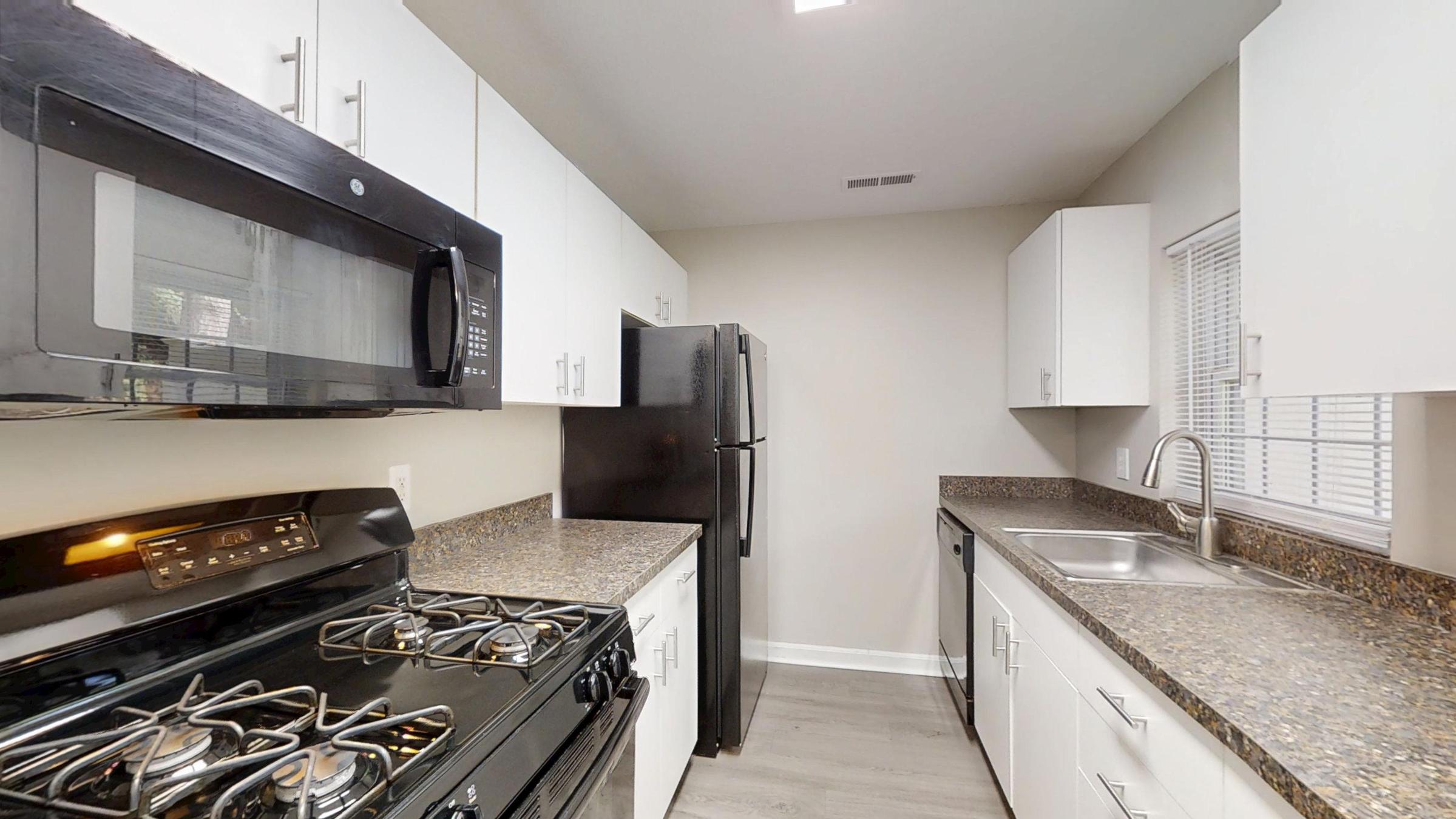 A modern kitchen featuring a black gas stove, a microwave, and a black refrigerator. The countertops are made of granite, and the cabinets are white. A stainless steel sink is installed under a window, and the flooring is light-colored. The space is well-lit and organized.