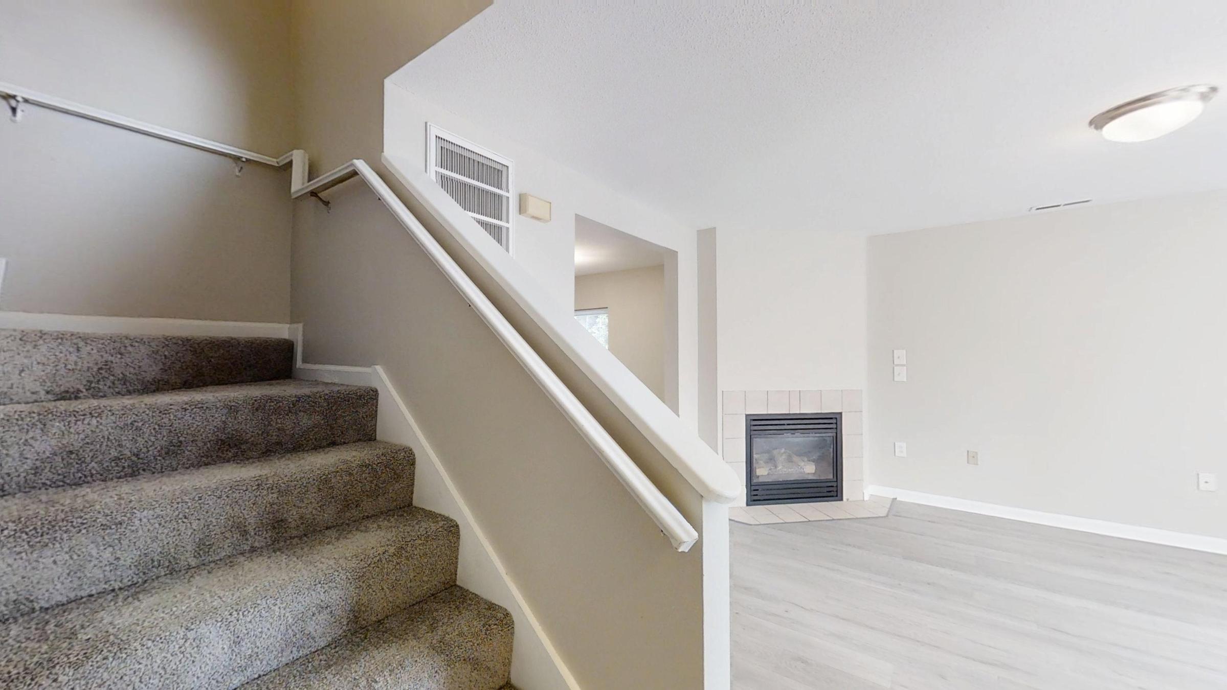 View of a staircase leading to an upper level, with beige carpet and a white handrail. To the right, there is a living area featuring light-colored walls and flooring, along with a fireplace in the corner. Natural light is coming in from nearby windows, creating a warm and inviting ambiance.