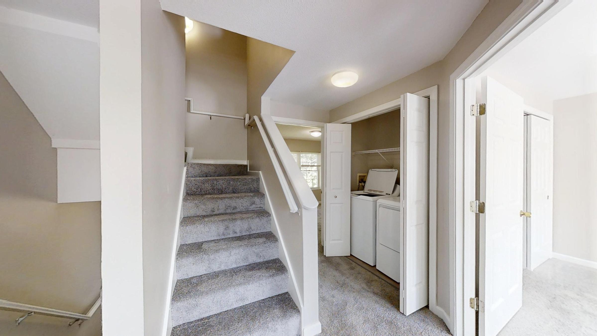 A light-filled hallway featuring a carpeted staircase leading to the upper floor. On the right, an open door reveals a laundry area with a washer and dryer. The walls are painted in a soft neutral color, and the space is well-organized, offering a warm and inviting atmosphere.