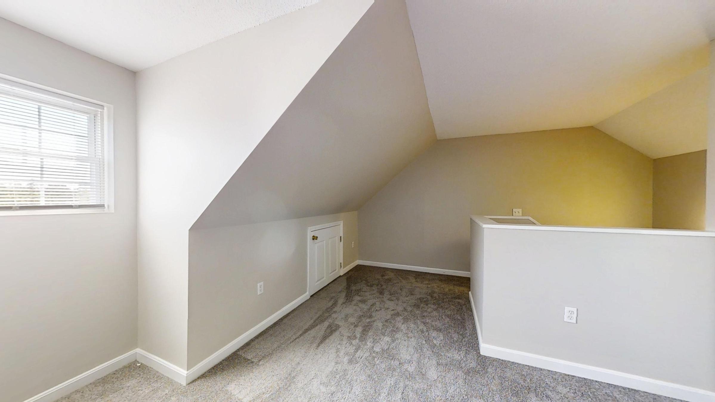 A cozy, partially finished attic space featuring sloped walls, light-colored walls, and carpeted flooring. A small window allows natural light to enter, and there is a heating unit on one wall. The area is open, with a railing visible at one end, suggesting a loft-like design.