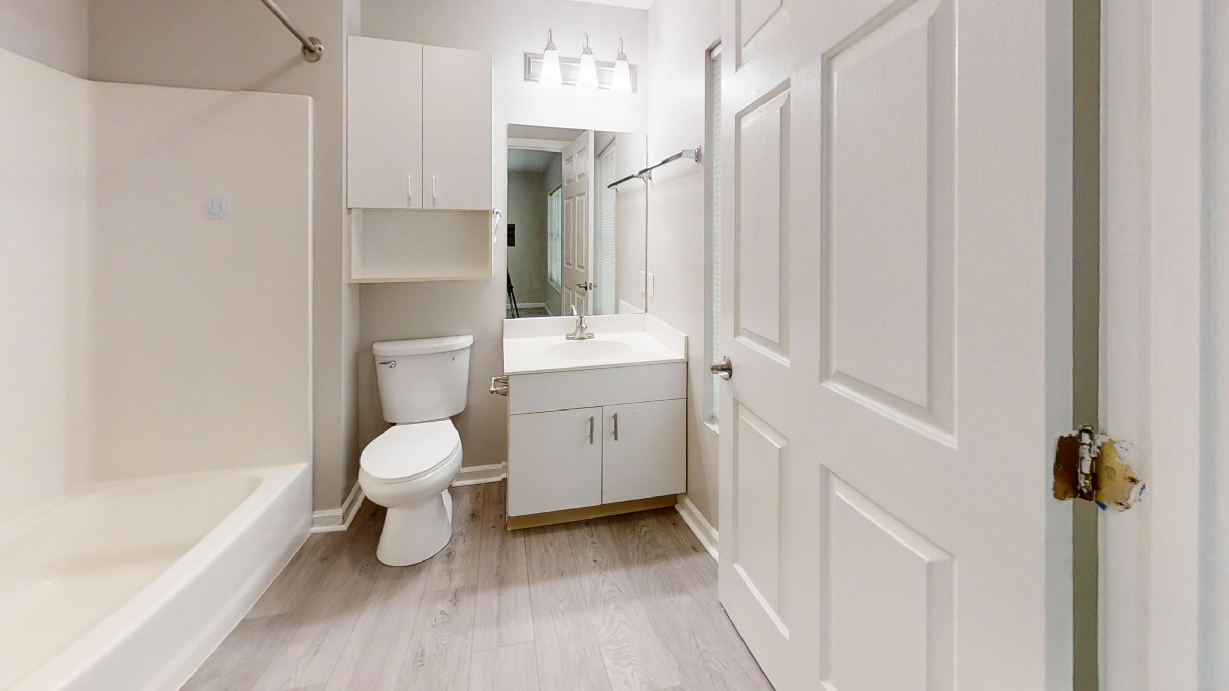 A clean and modern bathroom featuring a bathtub, toilet, and sink with a cabinet. The walls are painted in a light color, and there is a large mirror above the sink. The flooring is light wood. The bathroom door is partially open, leading to another area, and there are several light fixtures above the sink.