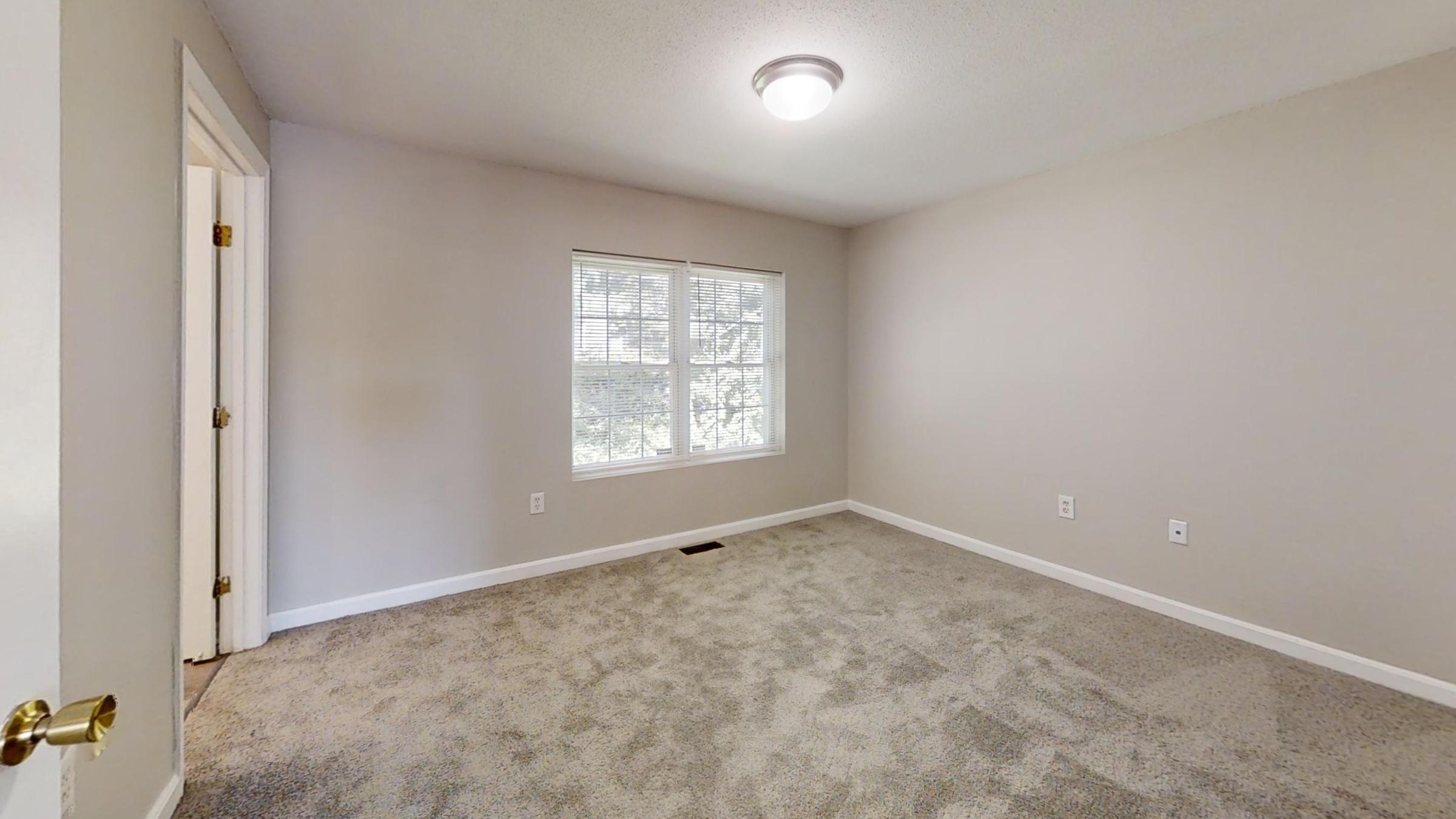A small, empty room with light gray walls and a carpeted floor. It features a window allowing natural light, a ceiling light fixture, and an open door leading to another space. The room appears clean and well-maintained, offering a simple and neutral design.