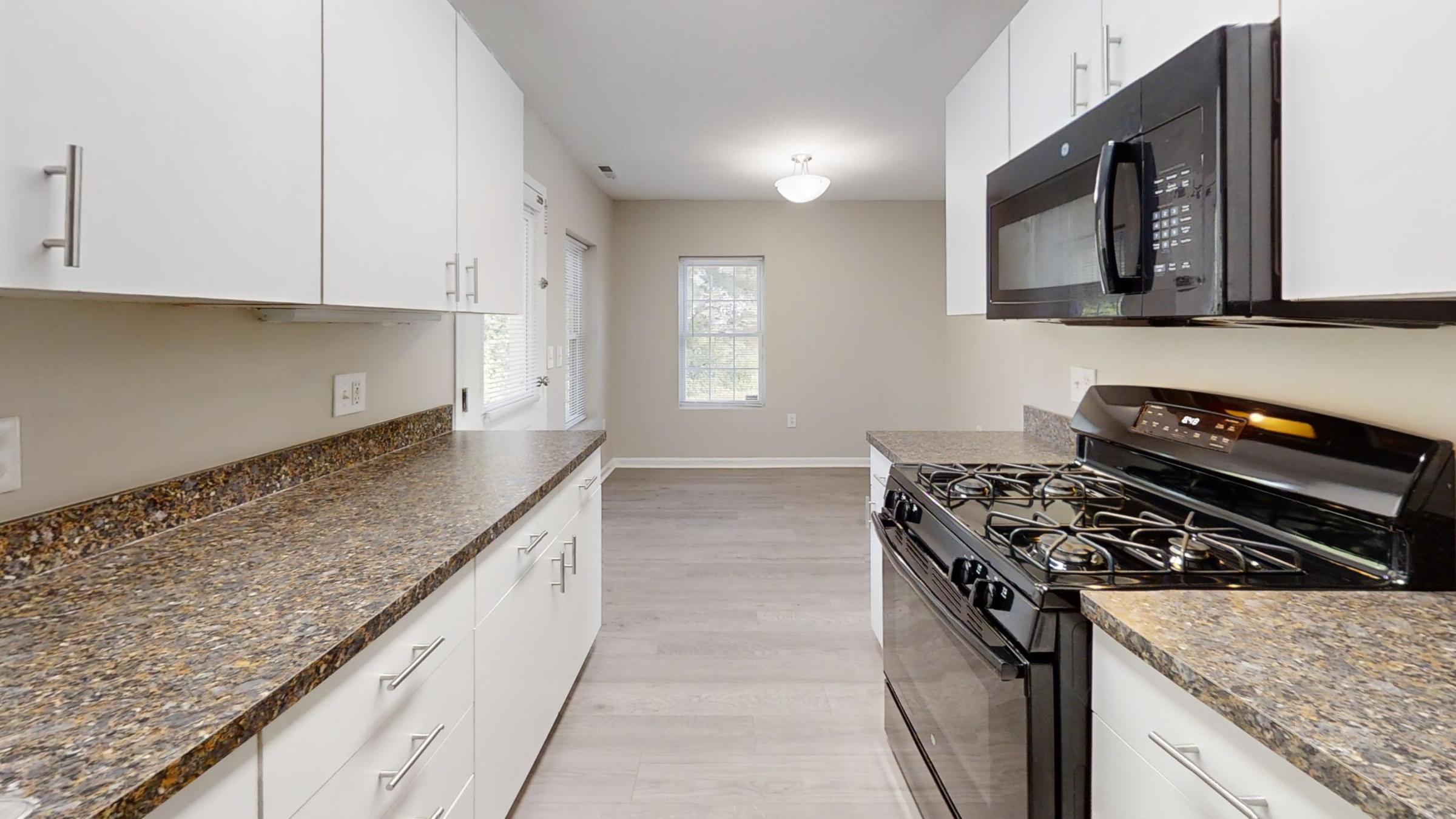 Modern kitchen with granite countertops, white cabinetry, and stainless steel appliances. A gas stove is on the left, and a microwave is above it. The space features a neutral wall color and a large window letting in natural light, creating an inviting atmosphere.