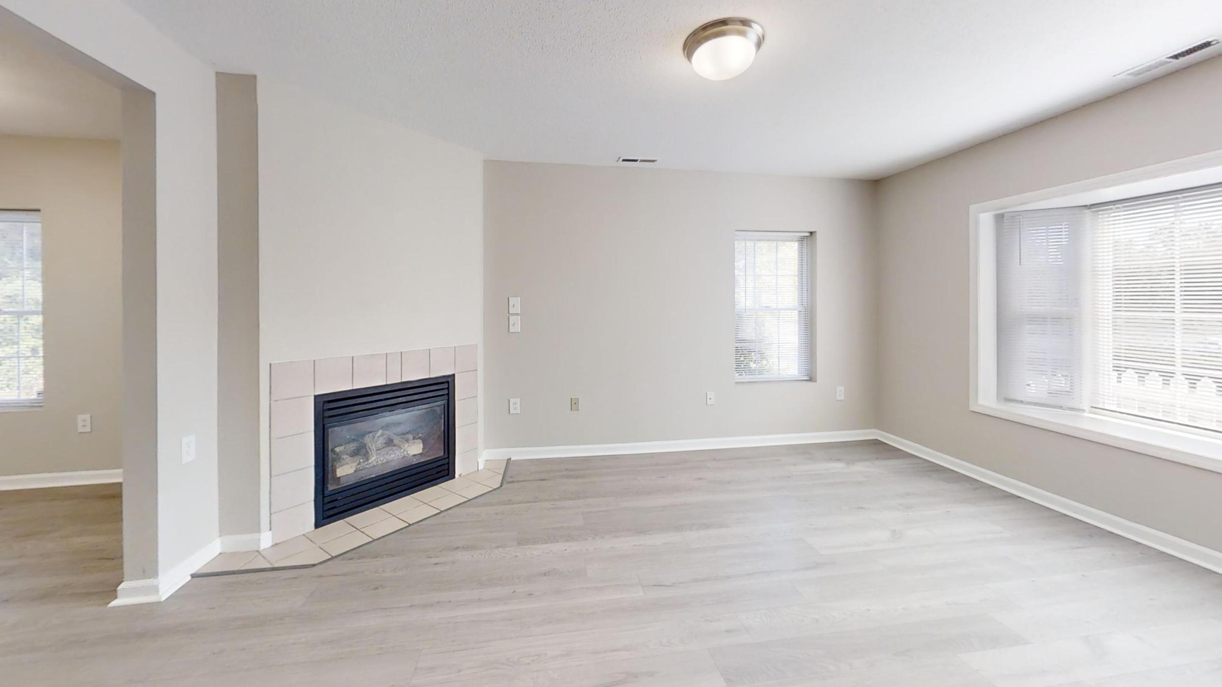 A spacious, empty room with light-colored walls and wood flooring. There is a fireplace with a tile surround on the left, and two windows allowing natural light. The room appears well-lit and open, suitable for various furniture arrangements.