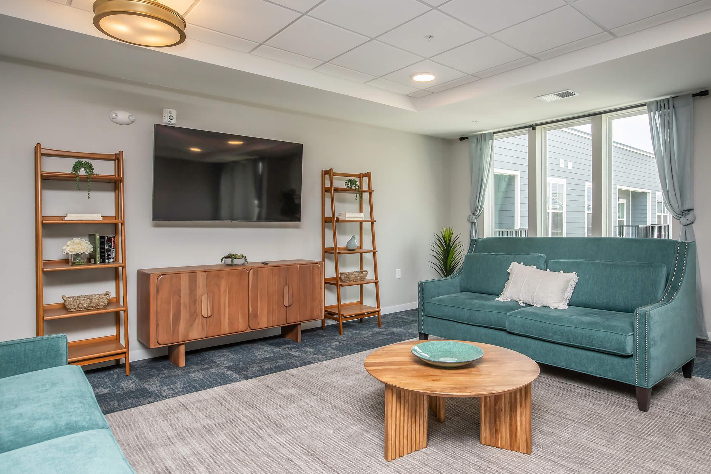 A modern living room featuring a teal couch, a wooden coffee table, and a wooden media cabinet. There are two shelves filled with decor items and a flat-screen TV mounted on the wall. Large windows provide natural light, and the space is decorated with a plant and light-colored curtains.