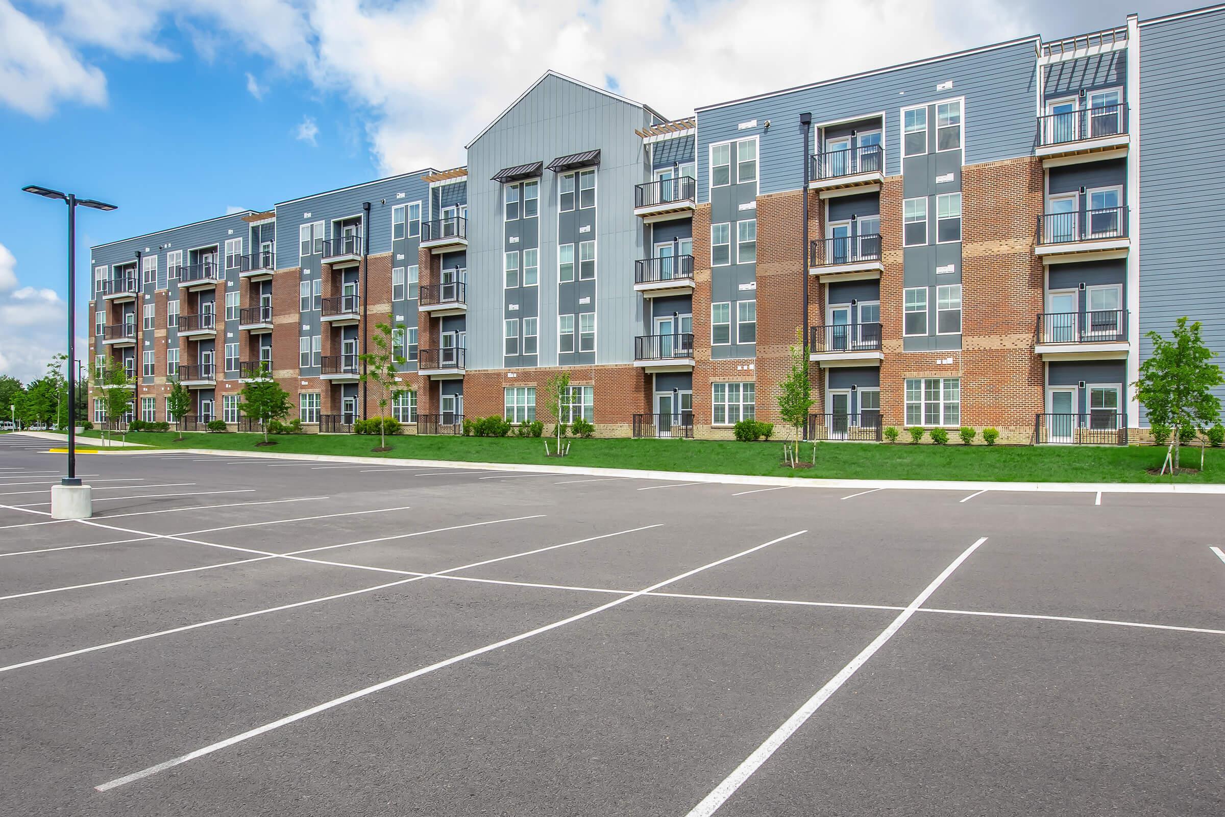 Modern apartment building with a mix of brick and gray siding, featuring multiple balconies. The foreground shows a large, empty parking lot with clearly marked parking spaces. Lush green grass and small trees are visible around the building, under a bright blue sky with scattered clouds.