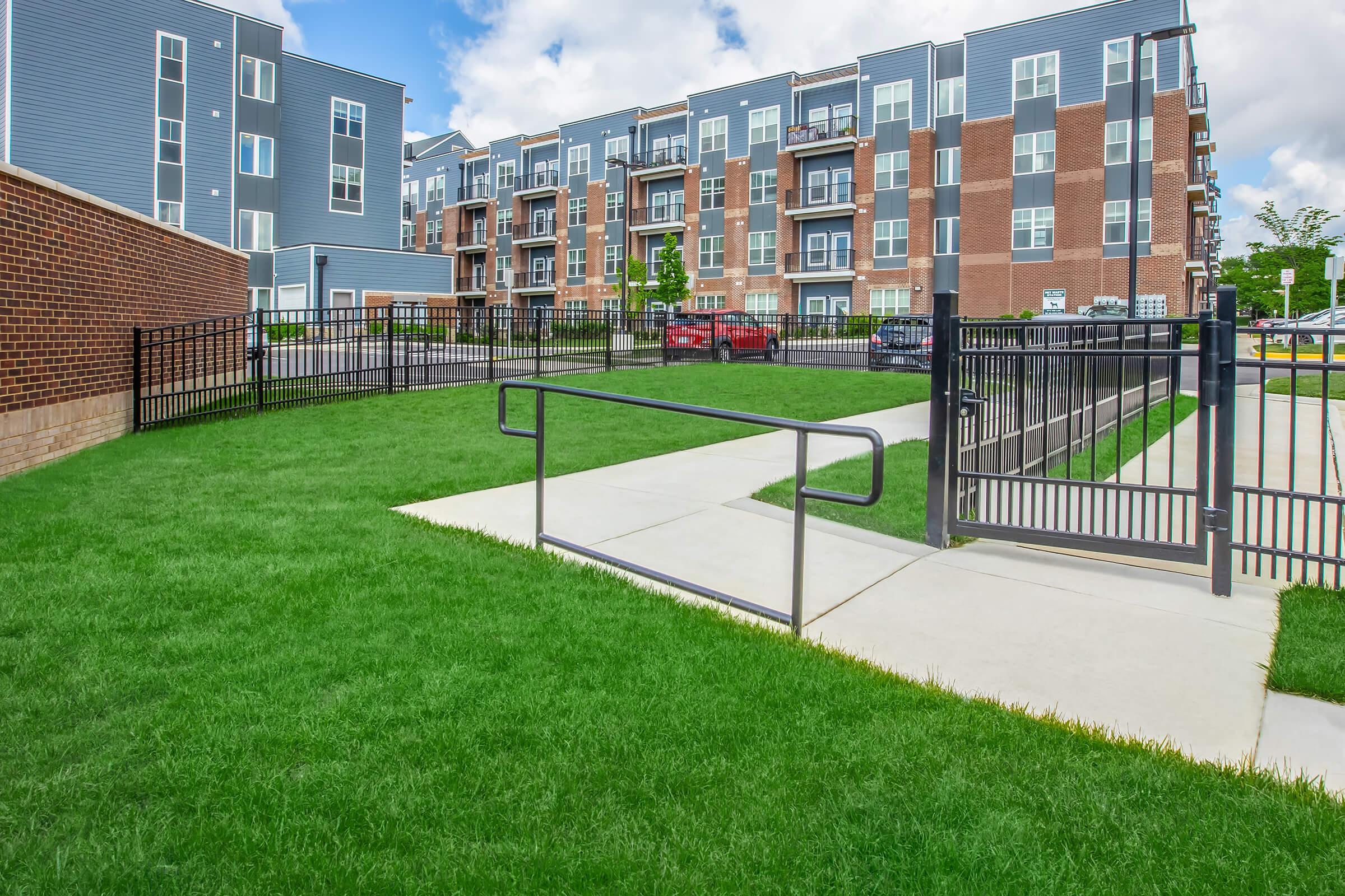 A view of a modern apartment complex with brick and blue siding. In the foreground, a smooth pathway leads through manicured green grass, bordered by black metal fences. The background features multiple apartment units with balconies and windows. The sky is partly cloudy, creating a bright, inviting atmosphere.