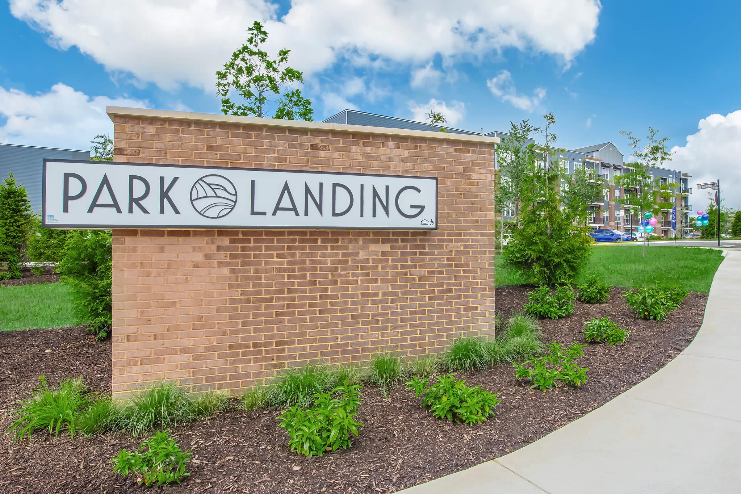 Sign for "Park Landing" mounted on a brick wall, surrounded by greenery and a pathway. The sky is blue with fluffy clouds, and the area has modern buildings in the background.