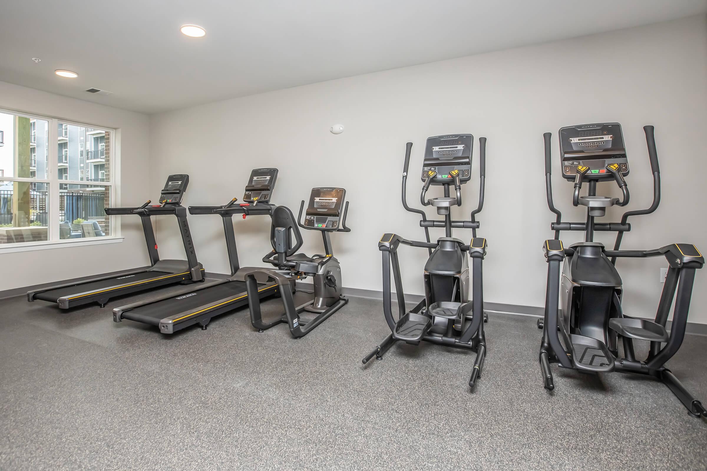 A modern gym space featuring four exercise machines, including two treadmills, a recumbent bike, and an elliptical machine. The area is well-lit with large windows providing natural light, and the flooring is designed for fitness activities.
