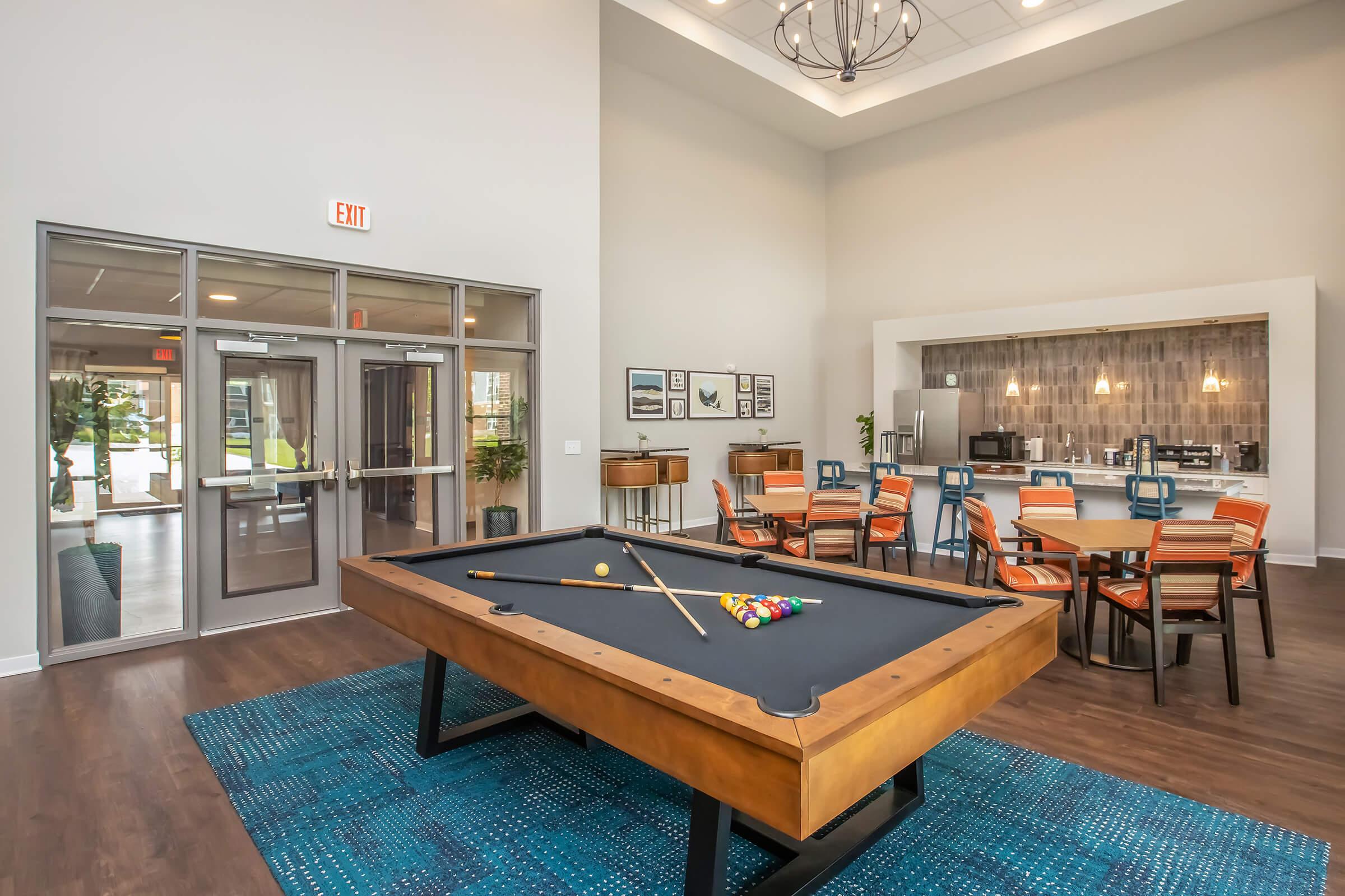A spacious indoor recreational area featuring a billiard table at the foreground, surrounded by a vibrant blue rug. In the background, there are several tables and chairs arranged for social gatherings, along with a kitchenette area. Large windows allow natural light to illuminate the space.