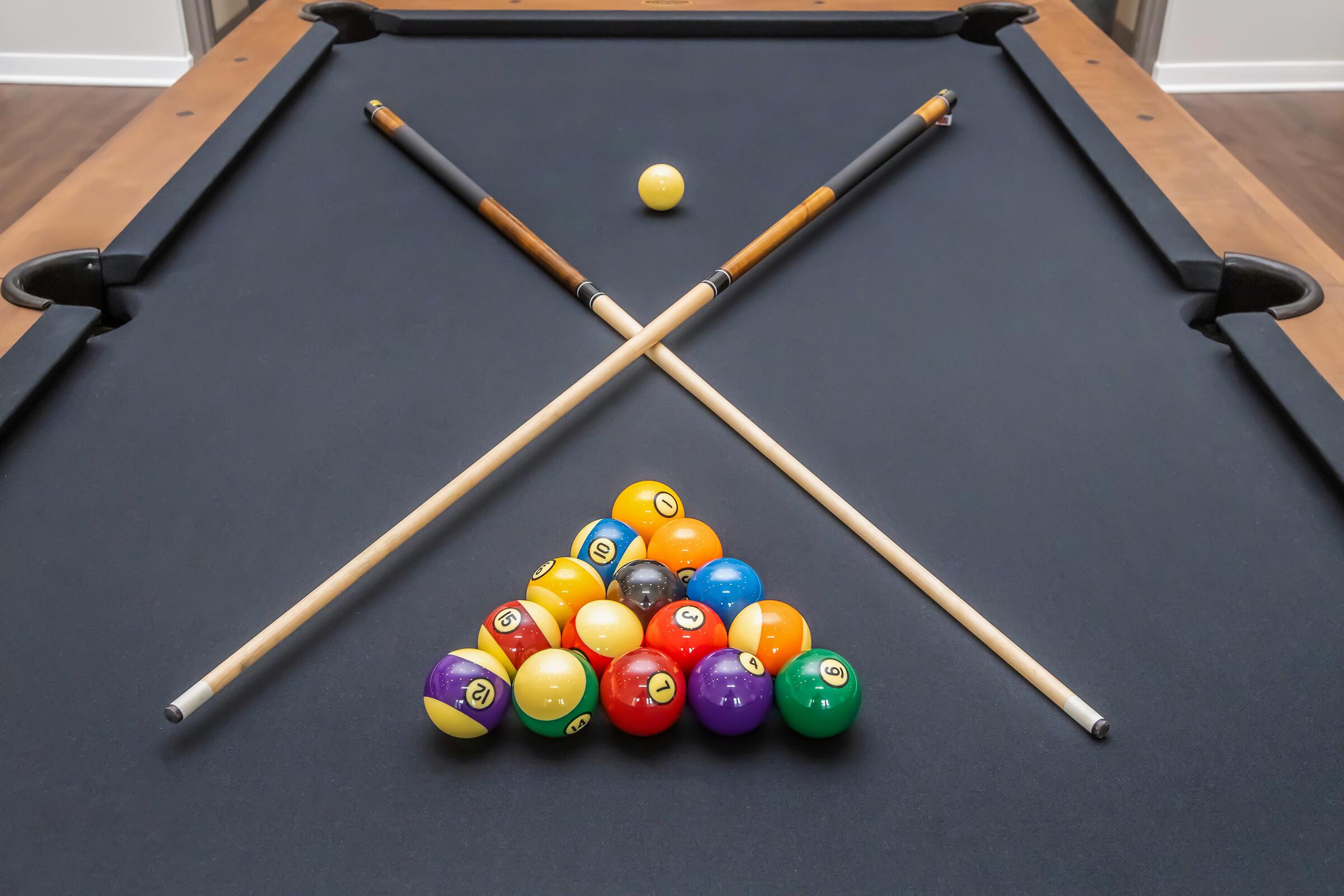A pool table with a triangle rack of colorful billiard balls and two crossed pool cues resting on the table's surface. A single cue ball is positioned on the table, ready for a game. The table has a dark felt surface and is set against a neutral backdrop.