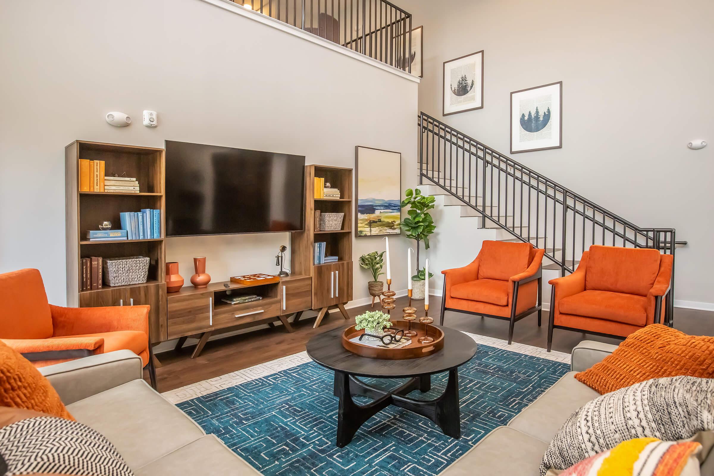 A modern living room featuring orange armchairs and a beige sofa, a round coffee table with decorative items, and a large flat-screen TV mounted on a stylish wooden media console. The space includes a patterned blue rug and artwork on the walls, with a staircase in the background.