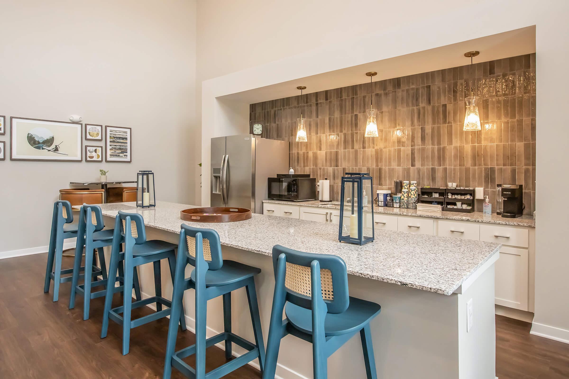 Modern kitchen bar with a granite countertop and eight blue high chairs. The wall features a stylish tiled backsplash with pendant lighting overhead. Various kitchen appliances, including a refrigerator and coffee machines, are visible in the background, creating a warm and inviting atmosphere.