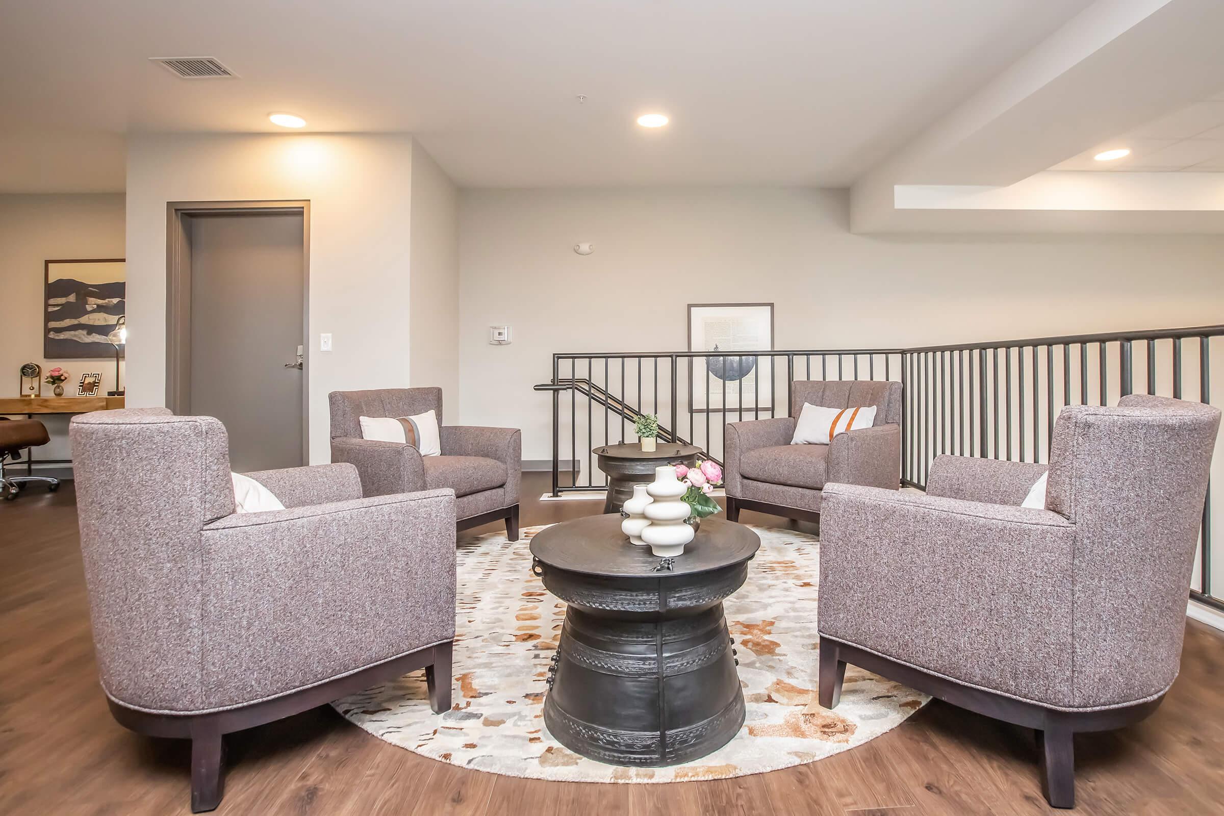A modern lounge area featuring four upholstered chairs arranged around a circular coffee table. The floor is wooden, and there's a round area rug with a subtle pattern. The walls are light-colored, and natural light enters the space, creating a welcoming atmosphere.
