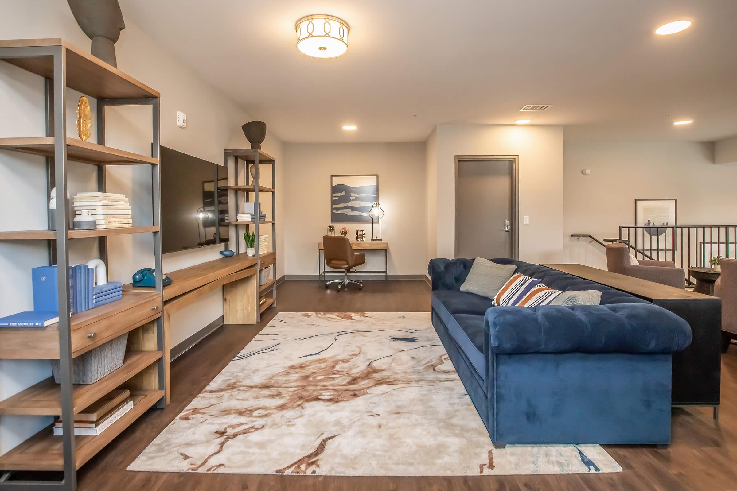 A modern living room featuring a navy blue couch, a patterned area rug, and a wooden shelf with decor and a TV. There’s a desk and chair in the background, with artwork on the walls. The space has neutral colors and a contemporary design, creating a cozy and inviting atmosphere.