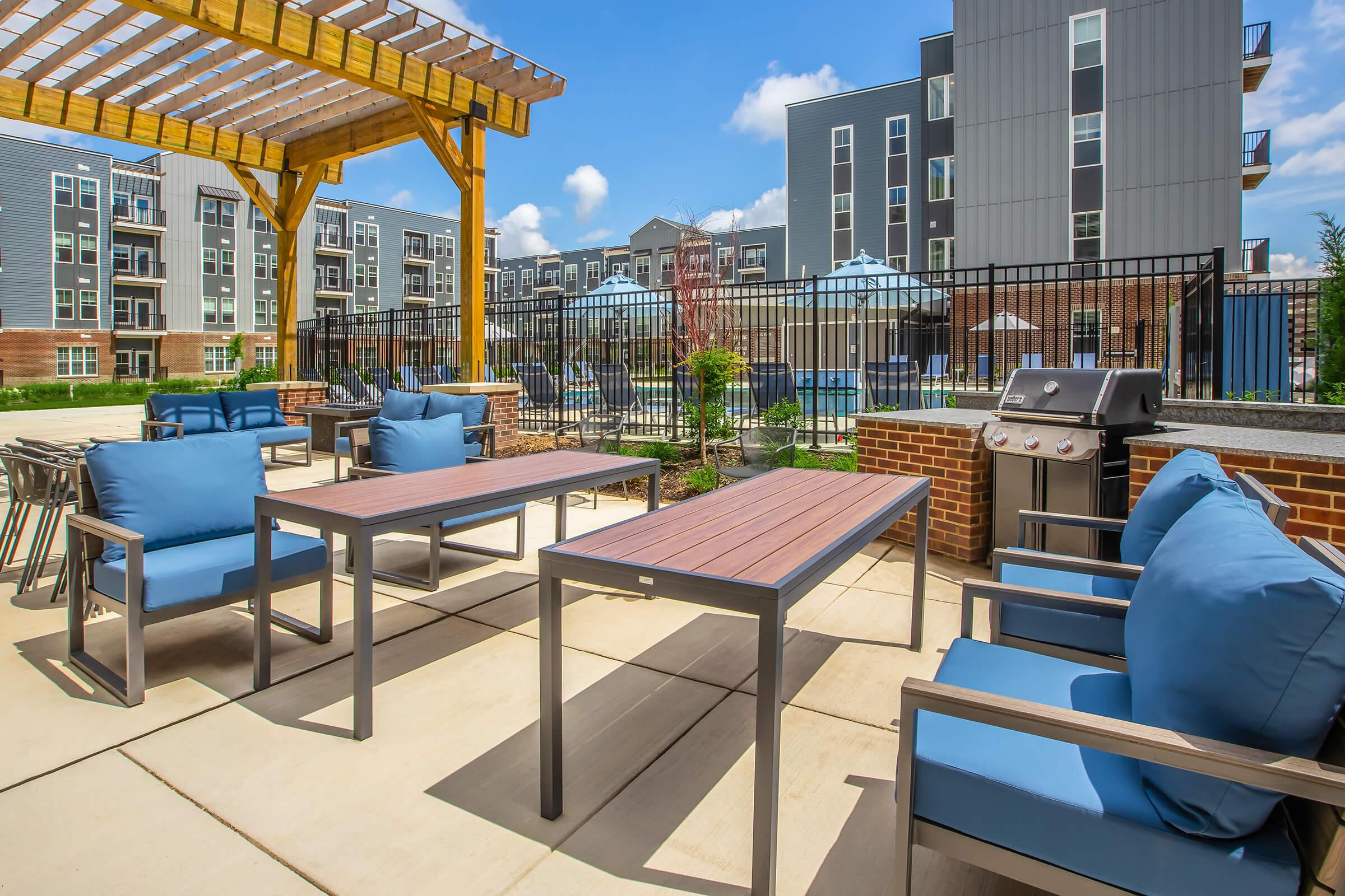 A modern outdoor patio area featuring blue cushioned chairs and tables, surrounded by a landscaped space. In the background, there are buildings and umbrellas providing shade. A grill is also visible, indicating a space for social gatherings and relaxation. The sky is bright and clear.