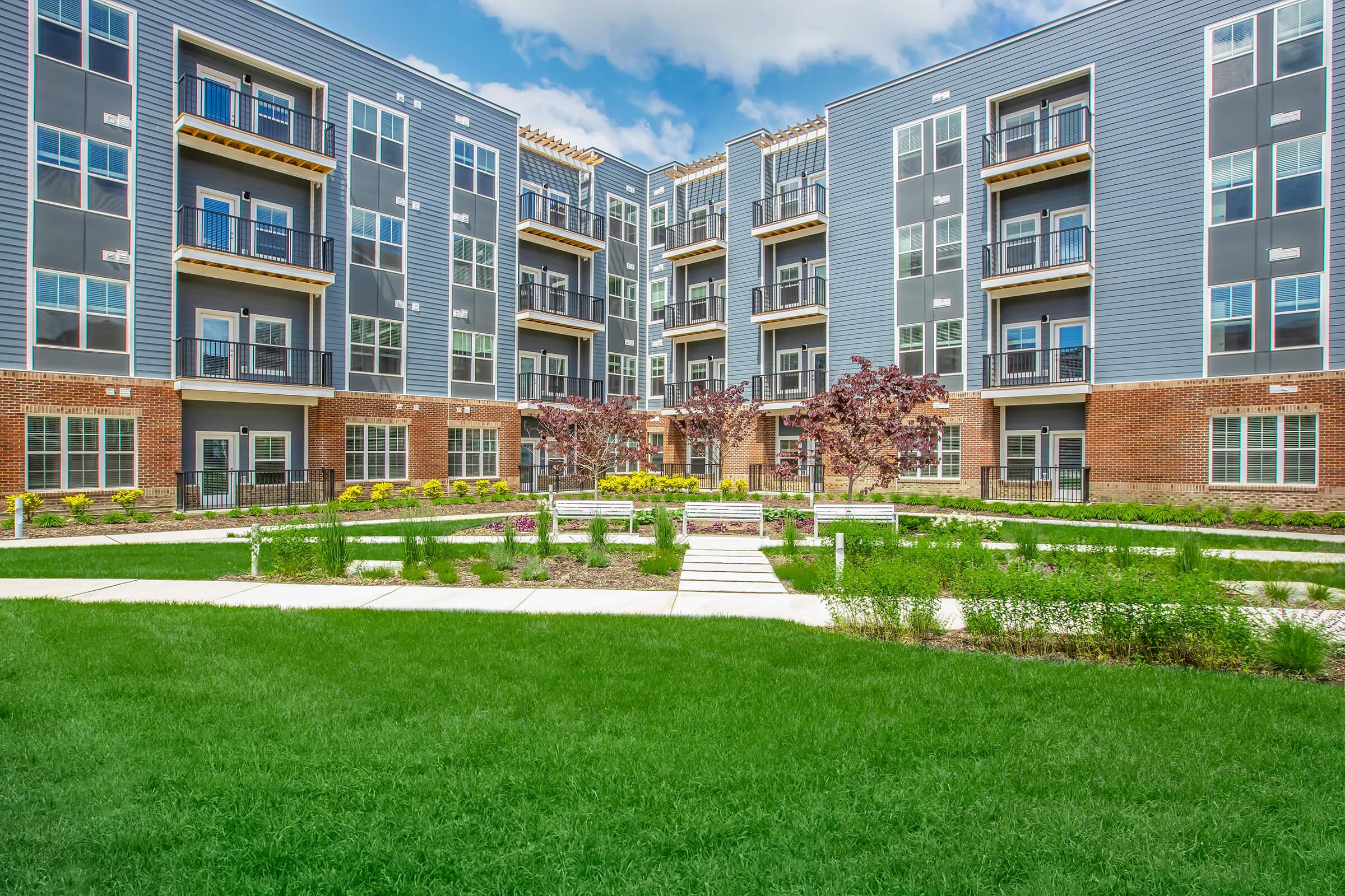 Modern apartment complex with multiple stories, featuring large windows and balconies. A well-maintained courtyard with green grass, flower beds, and pathways is visible in the foreground. The building has a contemporary design with a mix of gray and brick exteriors. Cloudy blue sky in the background.