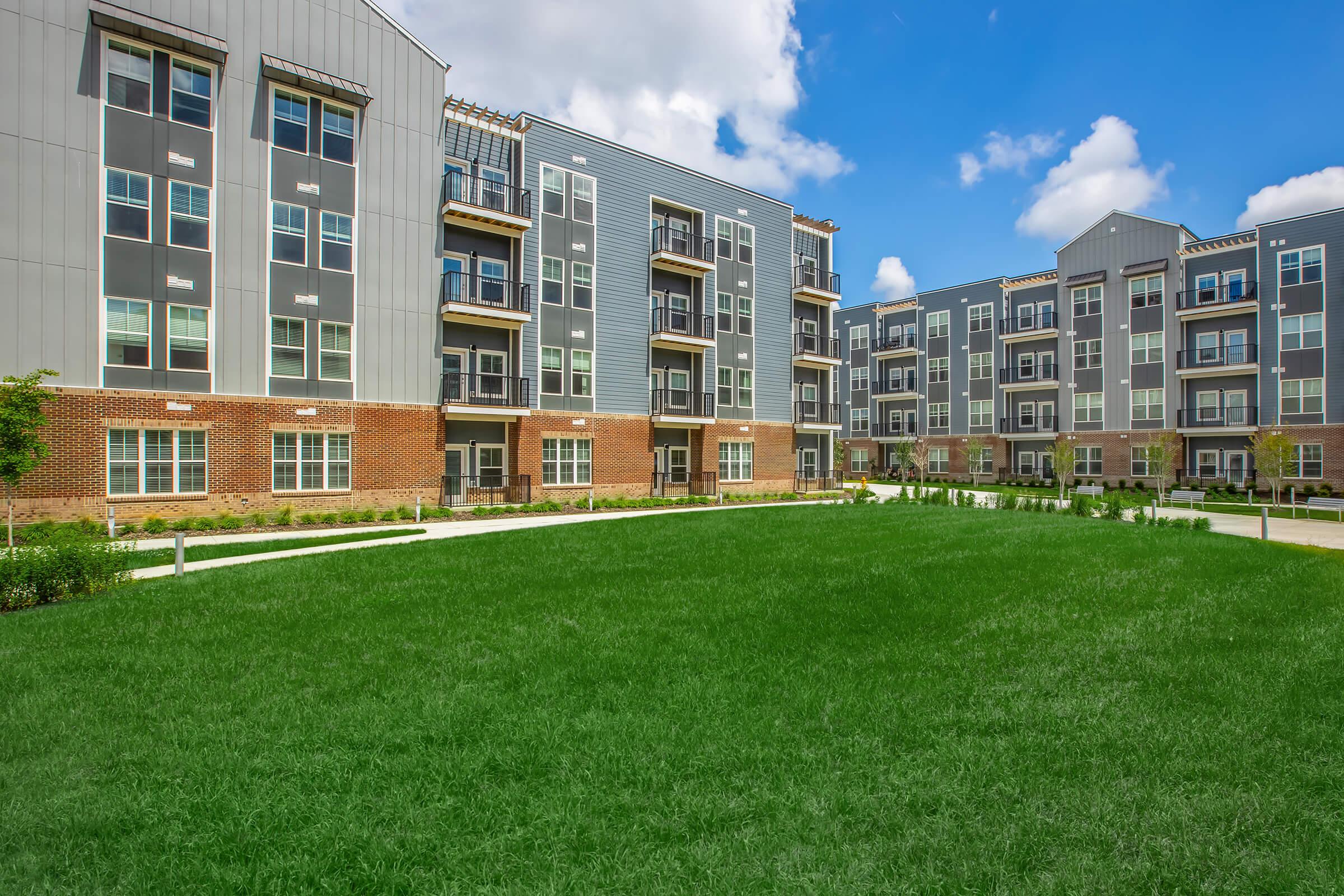 Modern apartment complex with gray and brick exteriors, featuring balconies and multiple stories. The foreground has a spacious, well-maintained green lawn, and the background shows additional buildings under a clear blue sky with scattered clouds.