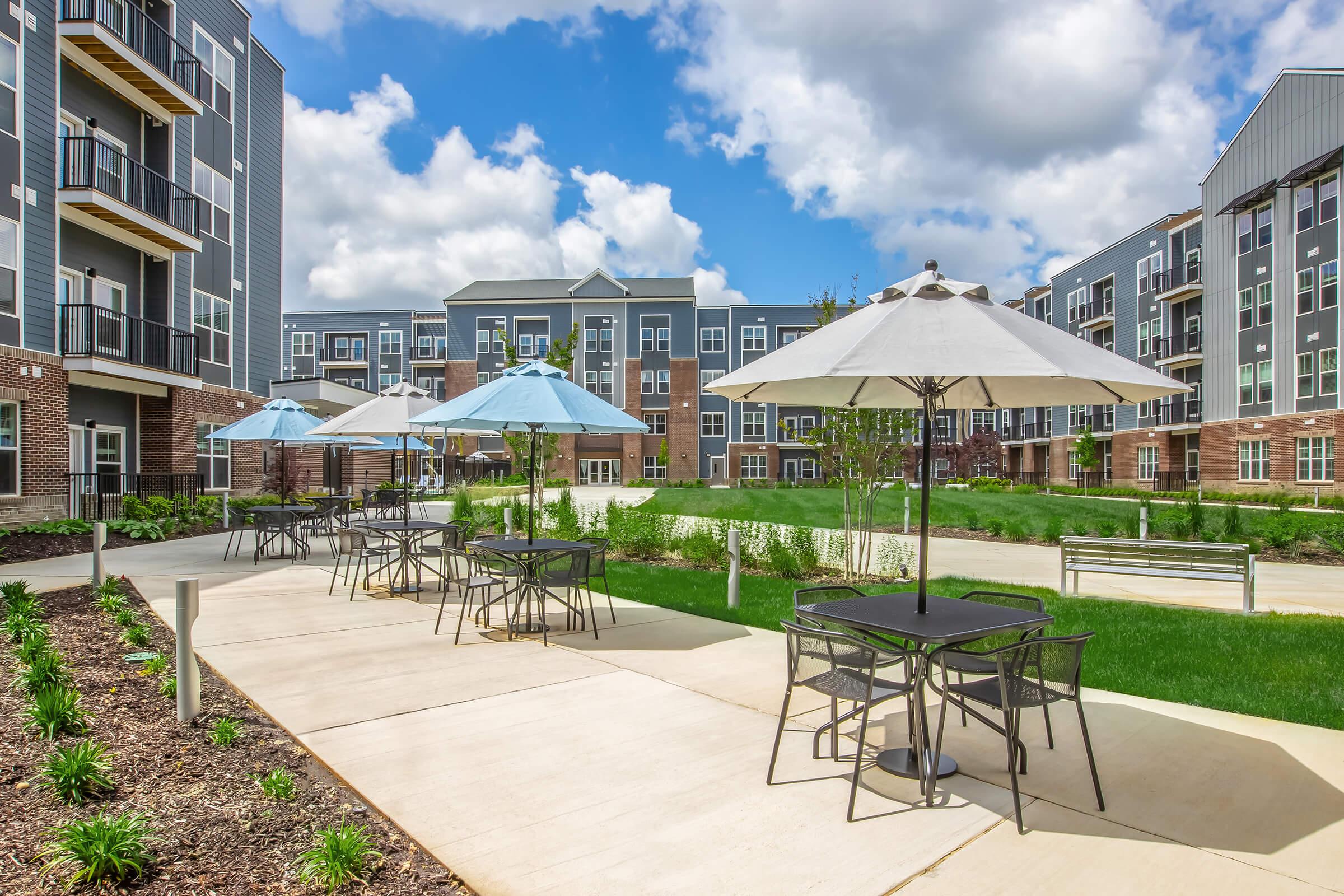 A modern outdoor space featuring a well-maintained courtyard with green grass, blooming plants, and multiple tables with umbrellas. Surrounding the area are contemporary apartment buildings under a bright blue sky with fluffy white clouds, creating a welcoming atmosphere for relaxation and socializing.
