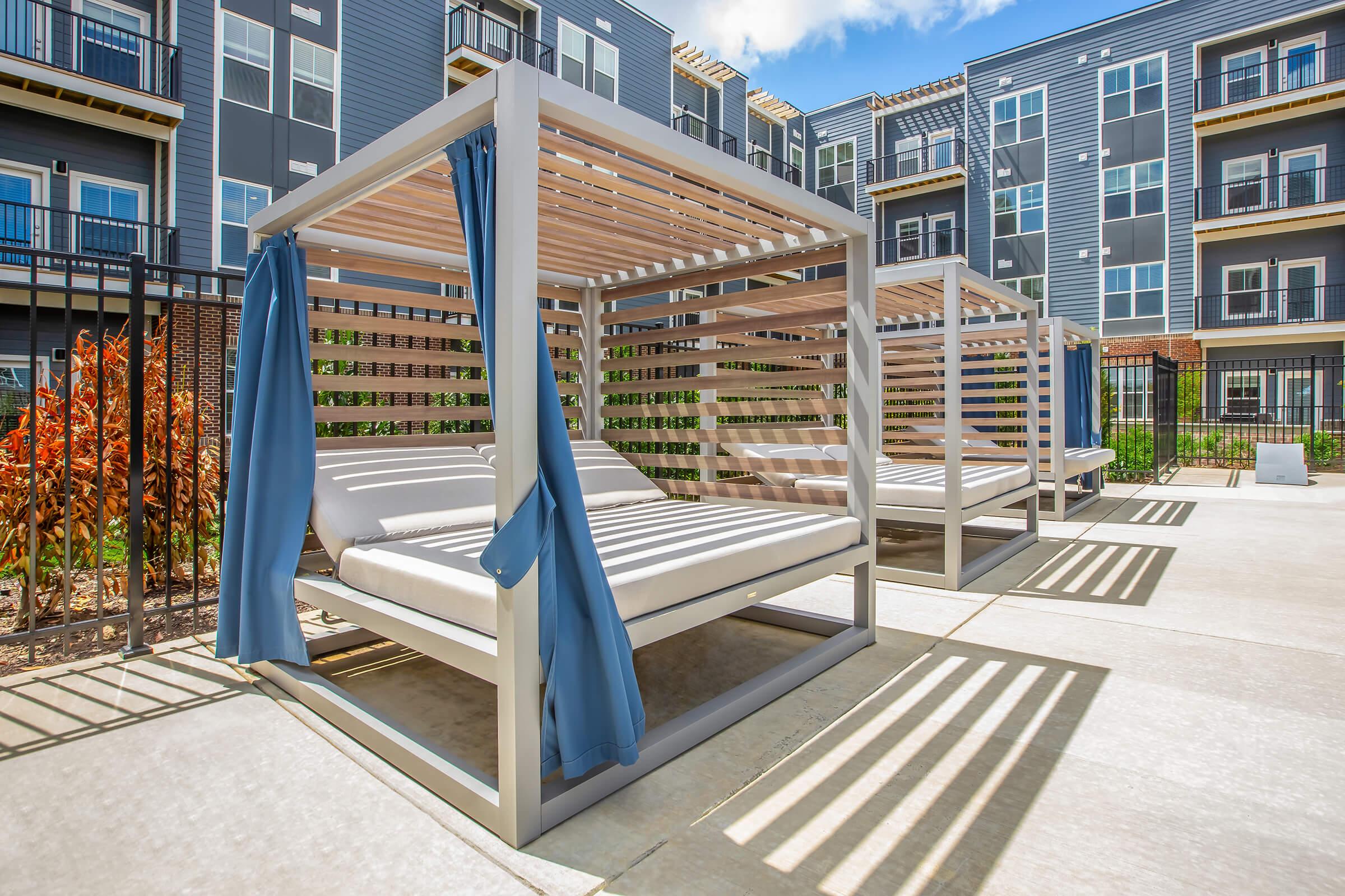 A modern outdoor lounge area featuring several shaded cabanas with wooden slats and blue fabric curtains. The cabanas are positioned on a concrete patio surrounded by greenery and modern apartment buildings in the background. Bright, sunny day with a clear blue sky.