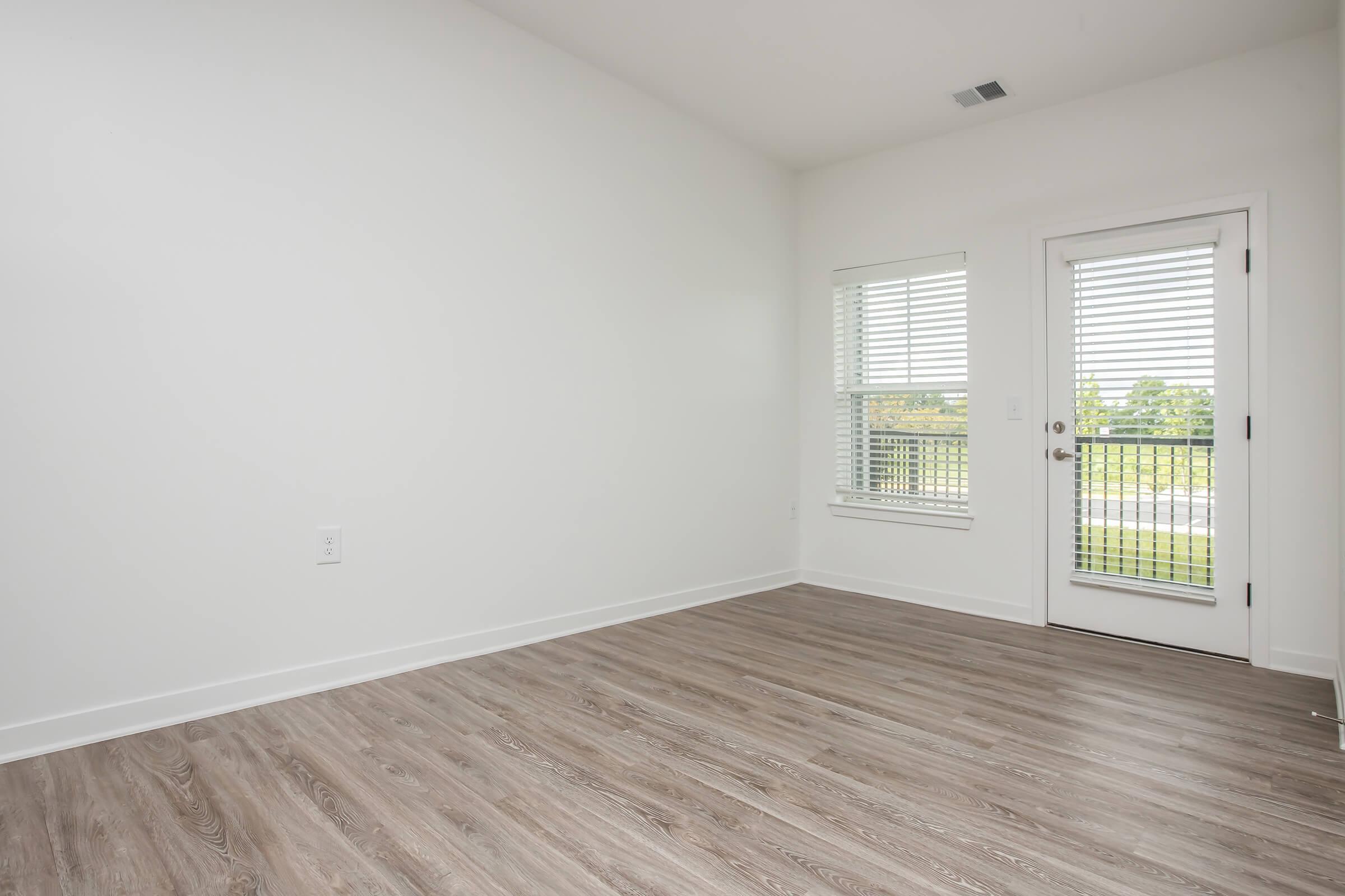 A spacious, empty room with light-colored walls and wooden flooring. There is a door leading to a balcony with blinds on a window. The room is well-lit, featuring natural light and a minimalist design.