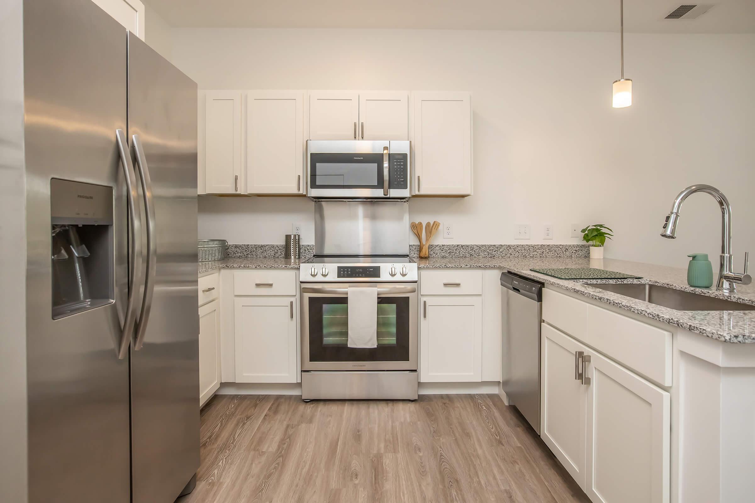 Modern kitchen featuring stainless steel appliances, including a refrigerator, microwave, and oven. The countertops are made of granite, and the cabinetry is white. A sink with a modern faucet is present, along with wooden utensils and a small plant for decoration. The flooring is a light wood look.