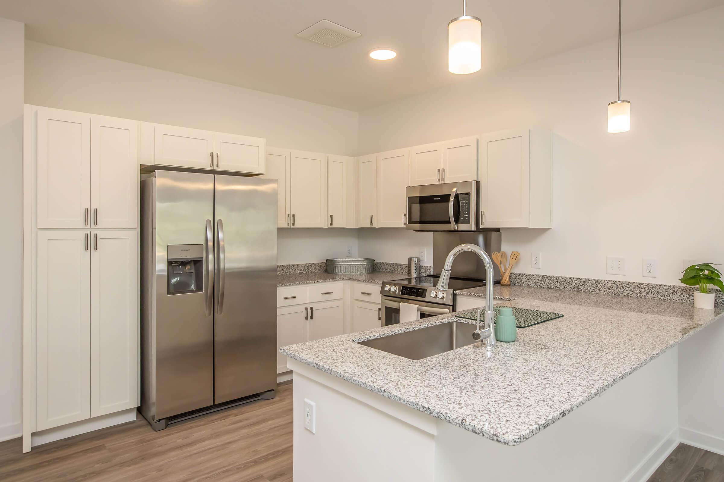 Modern kitchen featuring white cabinetry, a large silver refrigerator, and stainless steel appliances. The countertop is speckled gray and white, complementing the cabinets. There are pendant lights above, and a small plant adds a touch of greenery. A kitchen sink is integrated into the countertop.