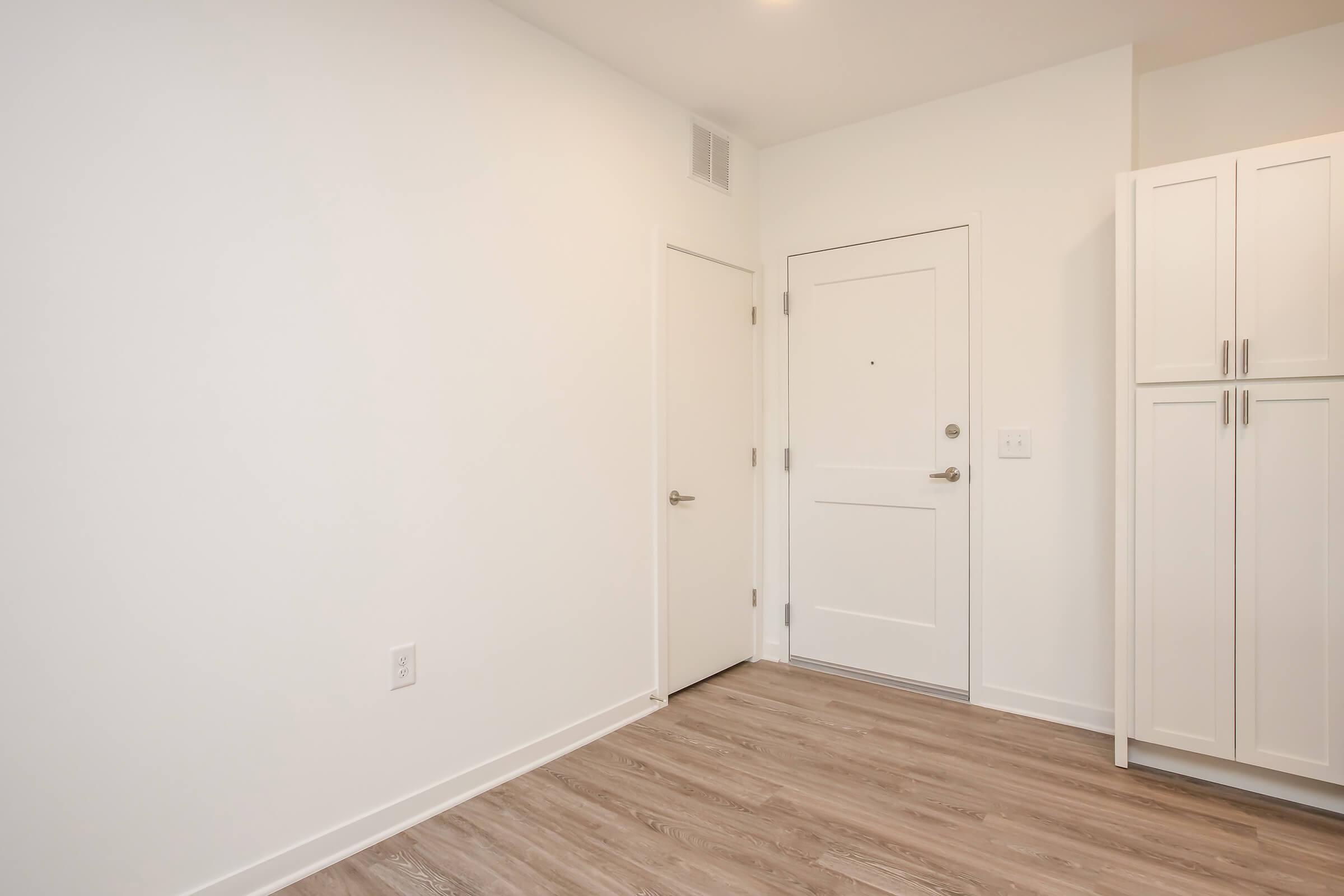 Interior of a minimalist room featuring light-colored walls, a wooden floor, a closed white door, and a front door with a small window. There is a white cabinet with multiple doors to the right, creating a clean and spacious feel.
