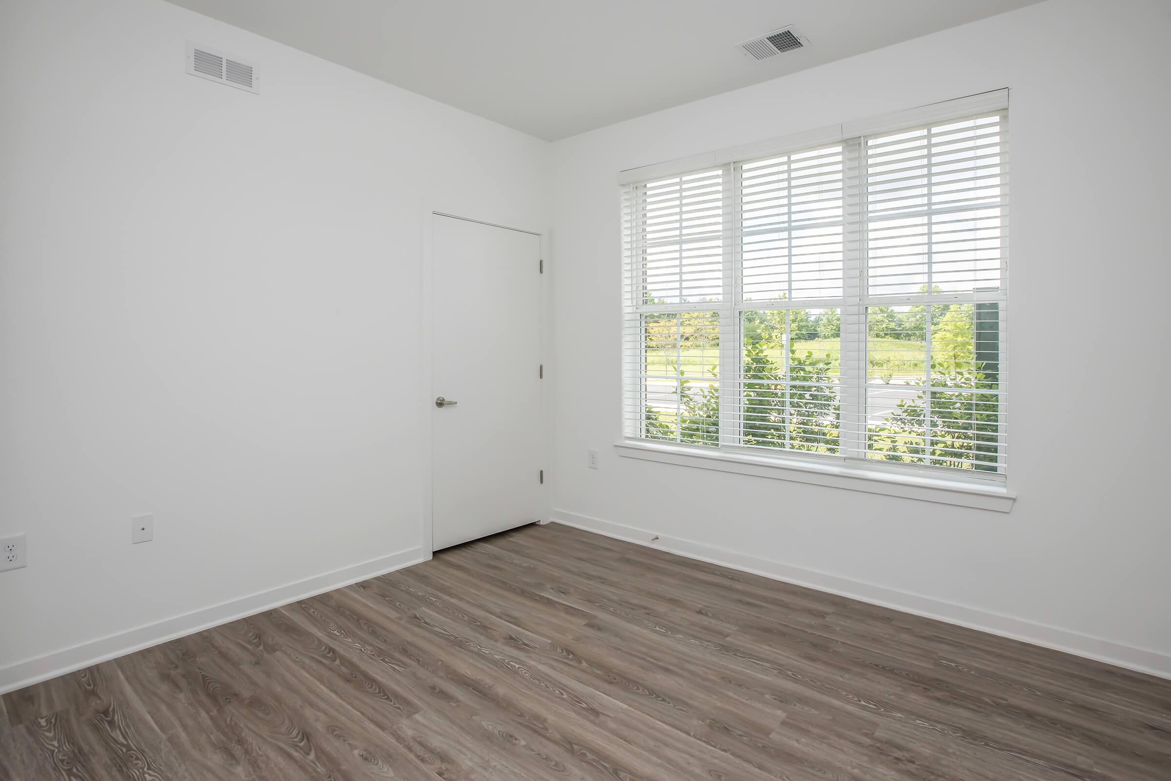 Empty room with light-colored walls and a wooden floor, featuring a large window with wooden blinds that offers a view of greenery outside. There is a door on one wall, and the room appears bright and spacious with natural light.