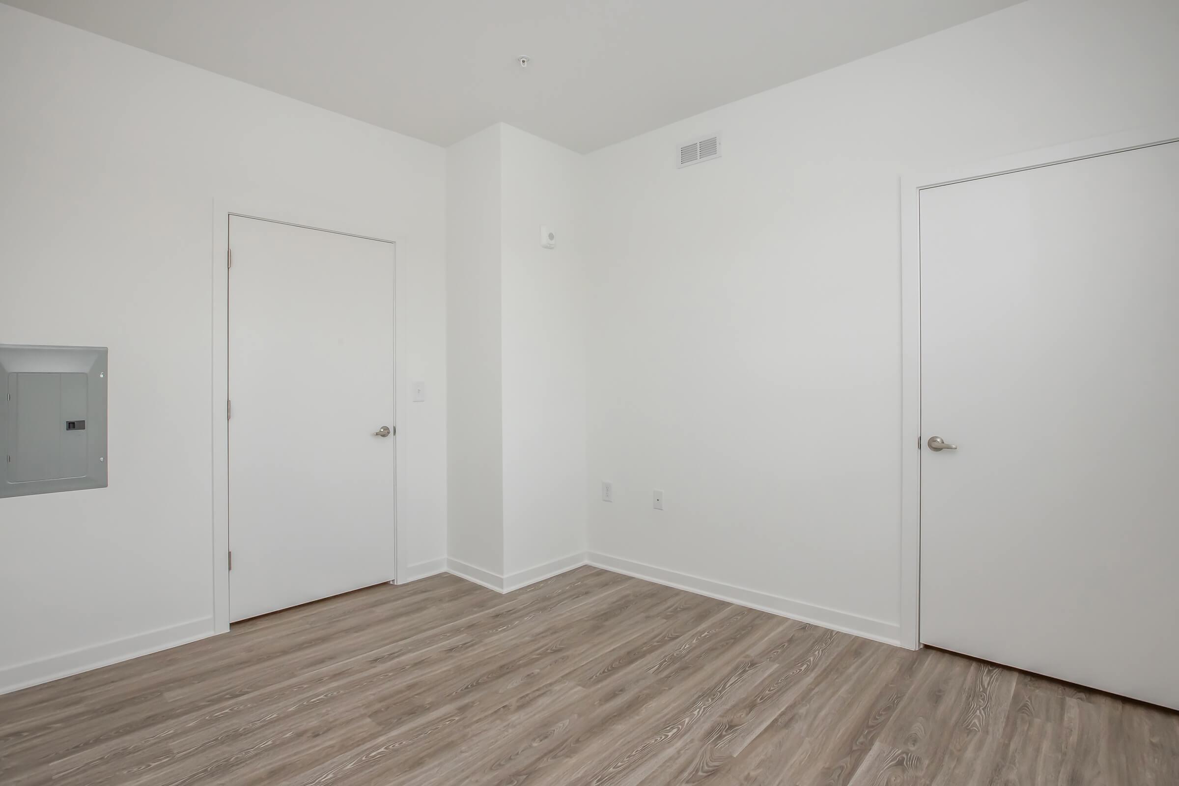 A minimalist interior of a room featuring light-colored walls and laminate flooring. There are two white doors, one leading to a closet or storage area and the other possibly to another room. A small electrical panel is mounted on the wall, and the overall space appears clean and modern.