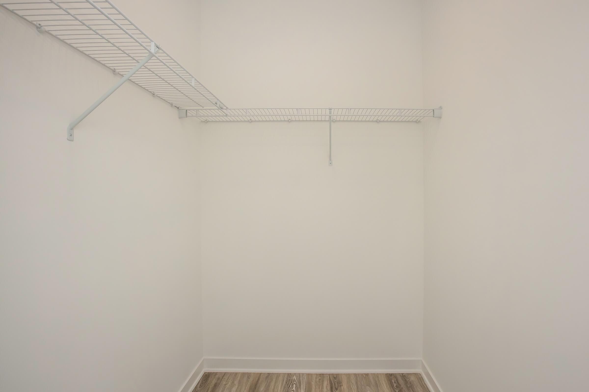 Empty closet with a light-colored wall, featuring a wire shelving unit on the left and a single horizontal wire shelf in the middle. The floor is covered with light-colored wood-like laminate. There are no items or additional furnishings present in the space.