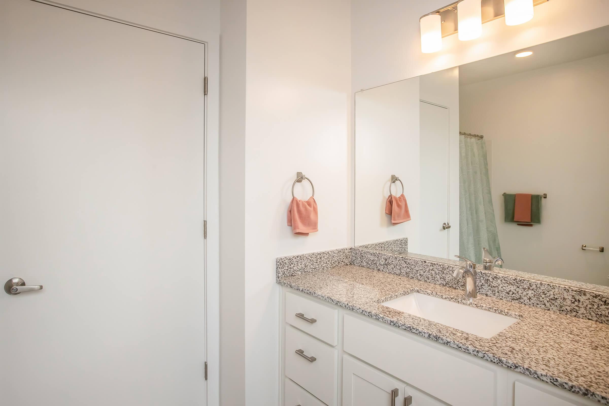 Modern bathroom featuring a clean white design. A large mirror is above a granite countertop with a sleek sink. Towel rings on the wall hold a pink hand towel, and a light fixture with three bulbs illuminates the space. A door leads to another area, and a light blue towel hangs nearby.