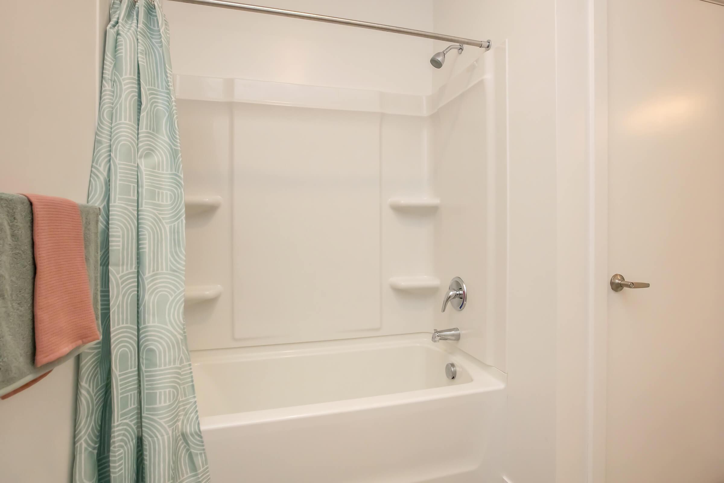 A clean and modern bathroom featuring a white bathtub with a showerhead, light teal patterned shower curtain, and minimal shelving. There are towel hangers with a light teal towel and a pink towel. The walls are painted white, creating a bright and airy feel.
