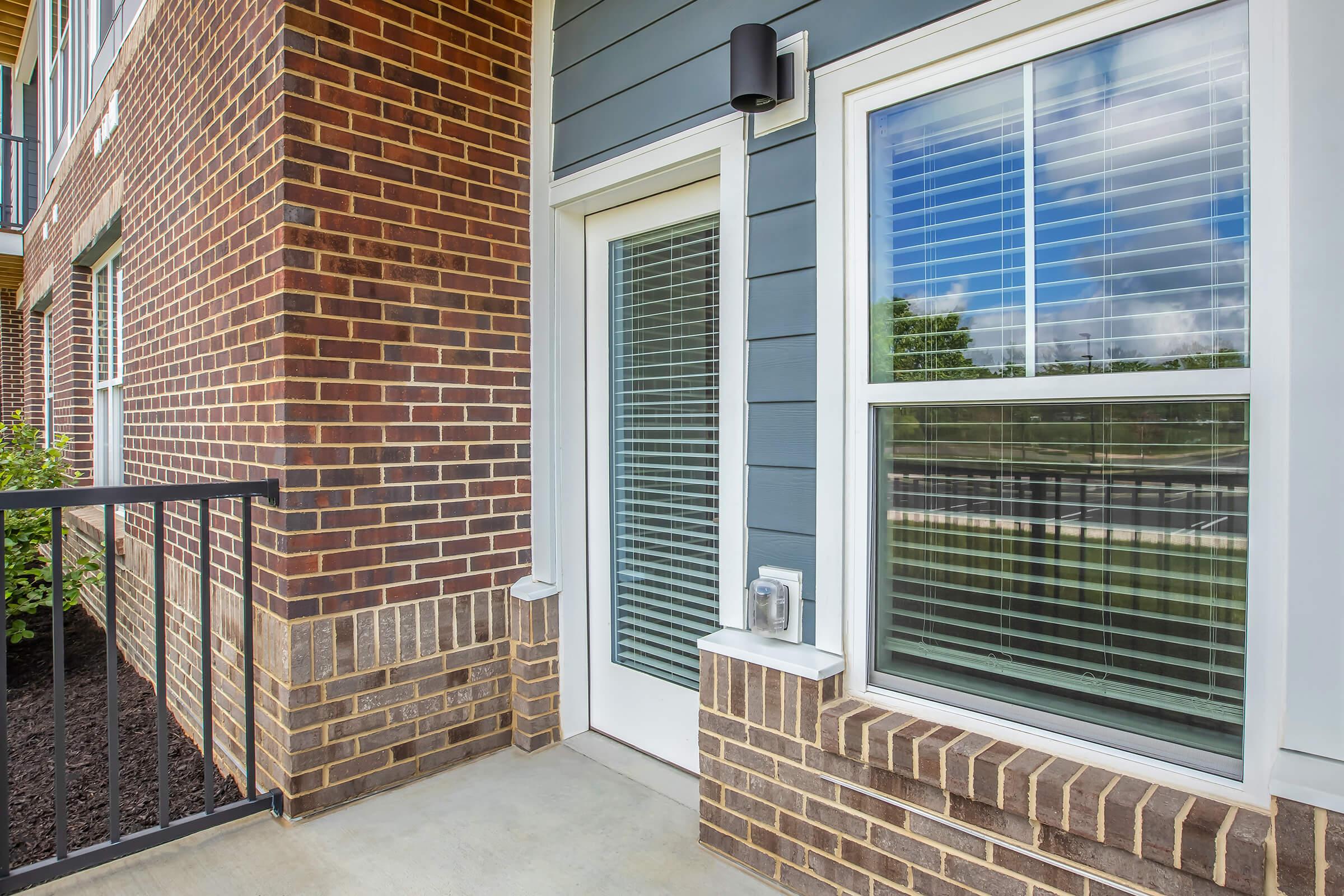 A close-up view of the entrance area of a modern building, featuring a glass door framed by large windows with white blinds, a brick exterior with a mix of brown and gray tones, and a small outdoor space with green grass visible in the background.