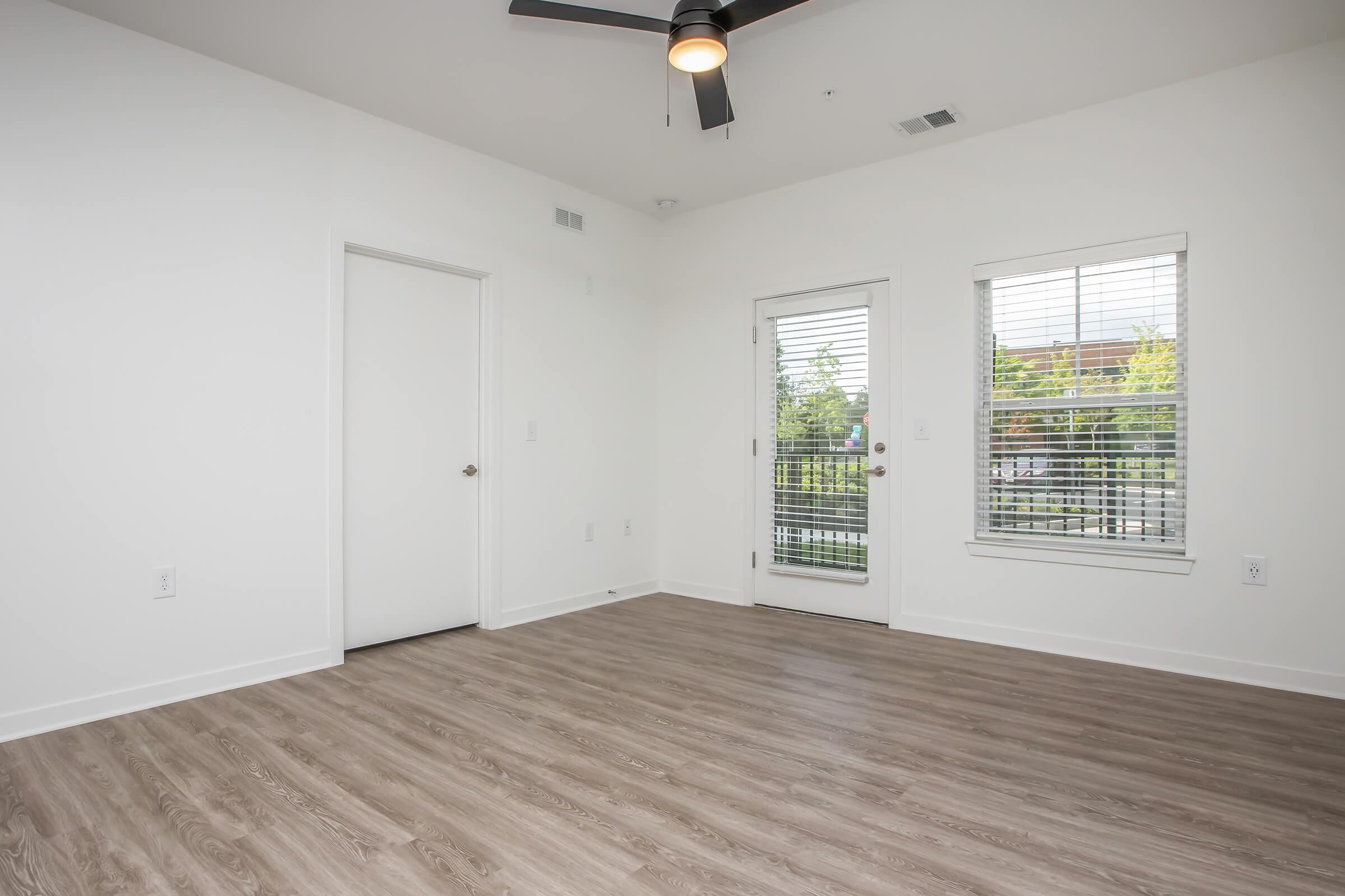 Bright, empty room featuring light-colored walls and a ceiling fan. It has a door leading to a balcony with large glass doors and windows, allowing natural light. The floor is made of light wood, creating a modern and spacious atmosphere.