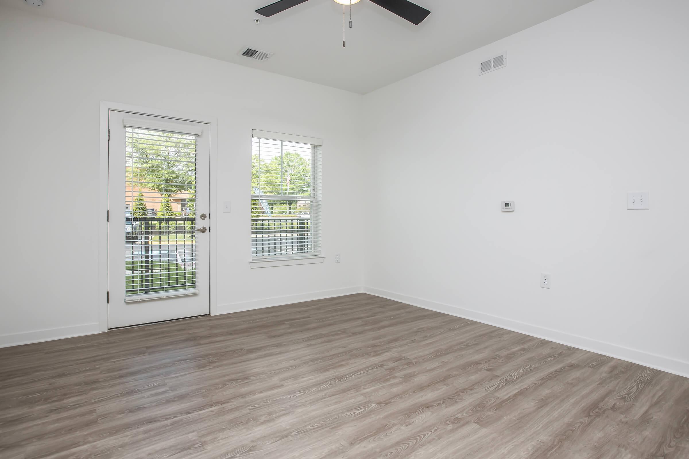 A bright, empty room with light-colored walls and a wooden floor. There is a ceiling fan and a glass door leading to a balcony, alongside a window with blinds. The space is designed for potential use as a living area or bedroom, with ample natural light and a minimalistic aesthetic.
