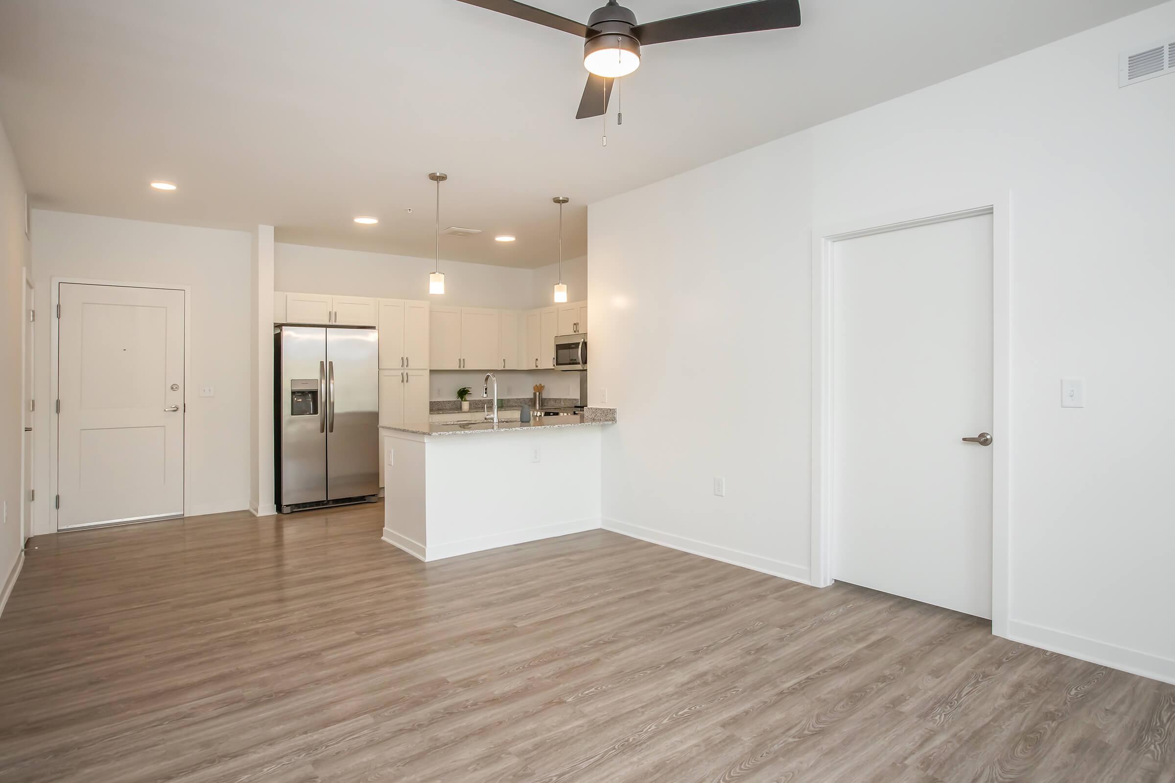 Modern open-concept apartment interior, featuring light wood flooring, a ceiling fan, and a kitchen with stainless steel appliances. The space includes a white wall, a door leading to another room, and is well-lit with pendant lighting.
